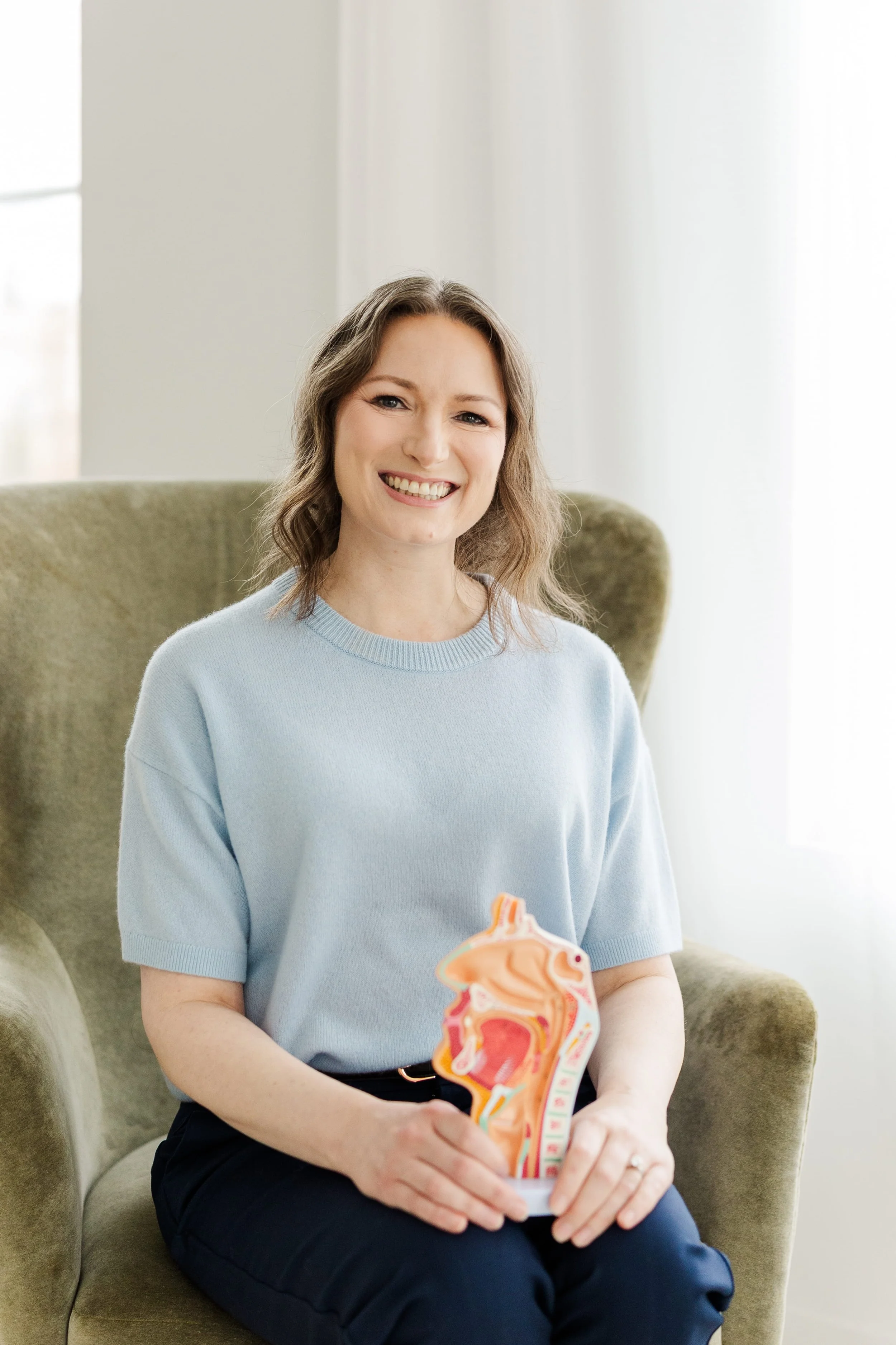 A smiling woman sitting in a green armchair is holding a detailed model of a human airway system. She is wearing a light blue sweater and navy pants, with a bright, well-lit room background.