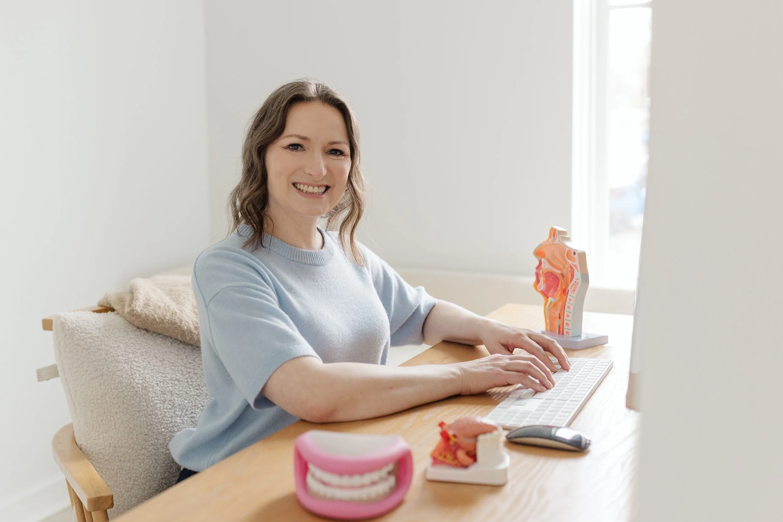 Woman smiling at the camera while working on a computer in a well-lit room decorated with anatomical models of human organs.