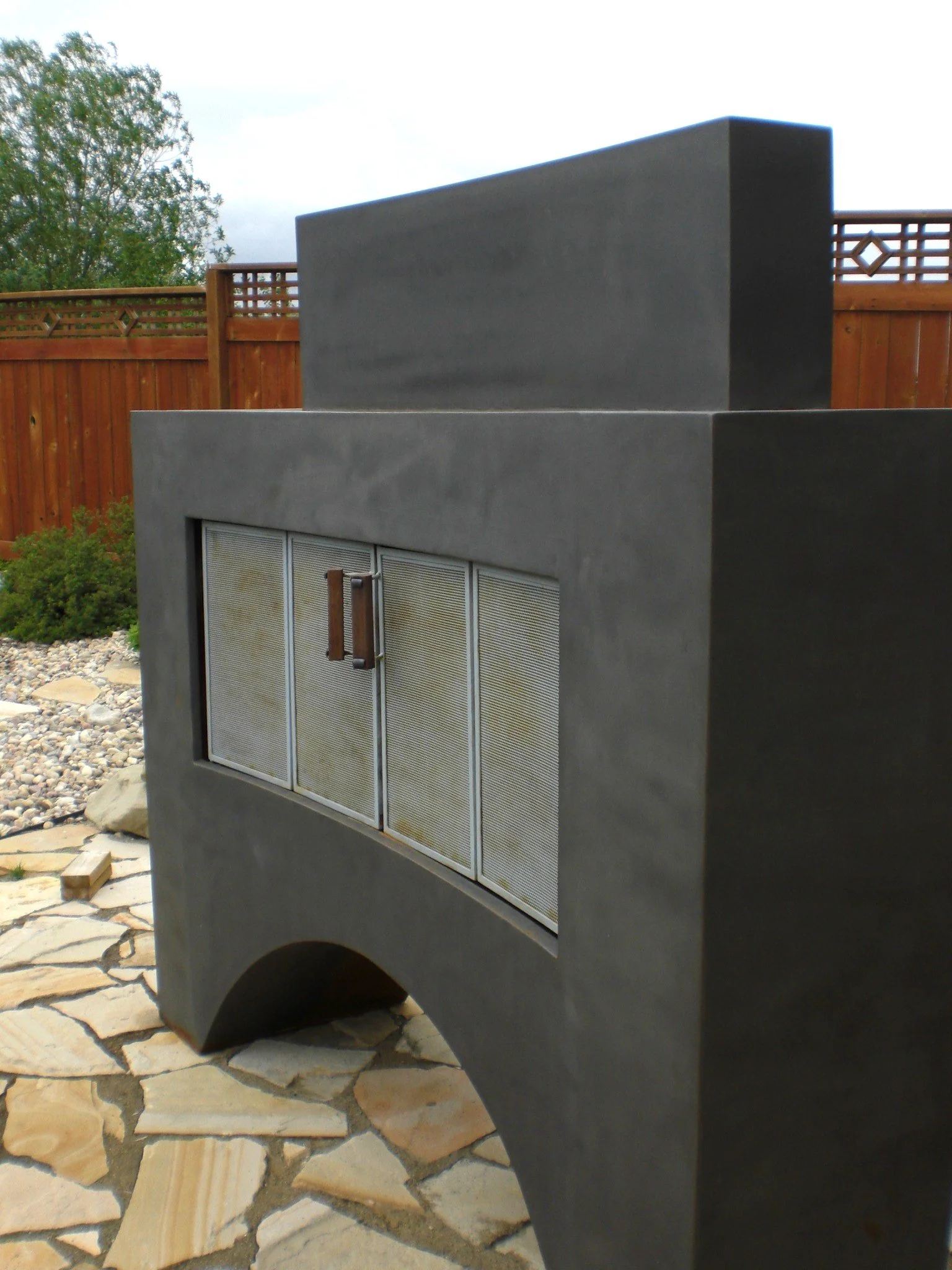 Close-up of modern outdoor fireplace with metal mesh doors and a curved stone base, set on stone patio with a wooden fence and trees in the background.