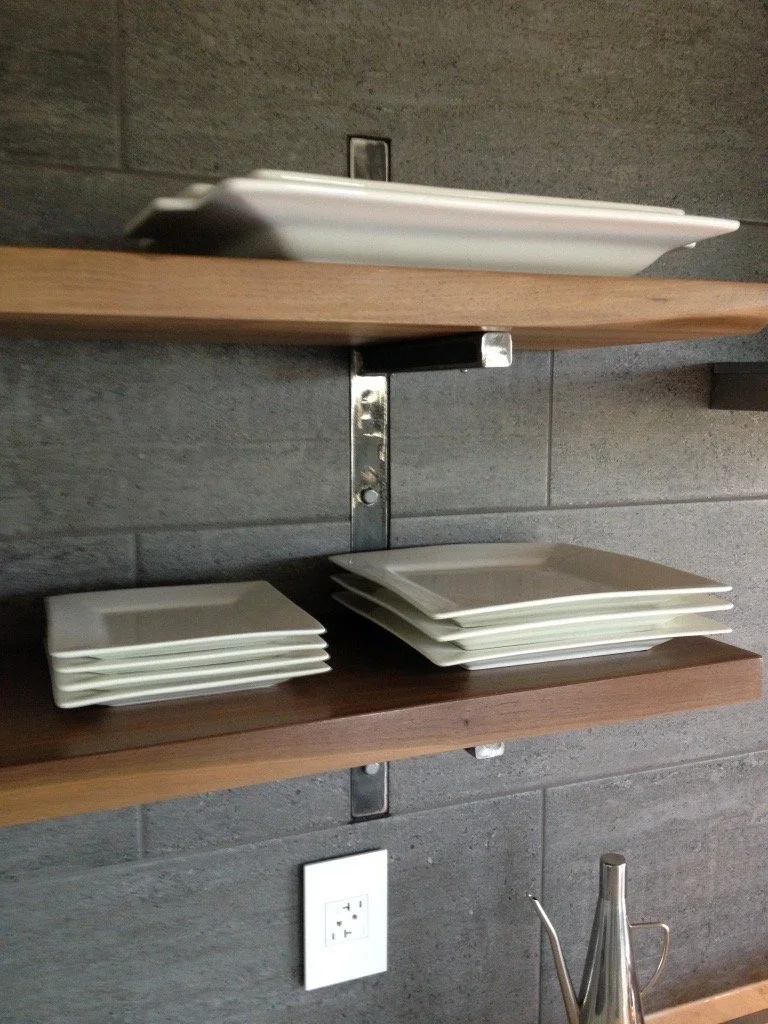 Three wooden shelves mounted on a gray wall with stacks of white plates on two of the shelves and a stainless steel teapot on the bottom right corner.