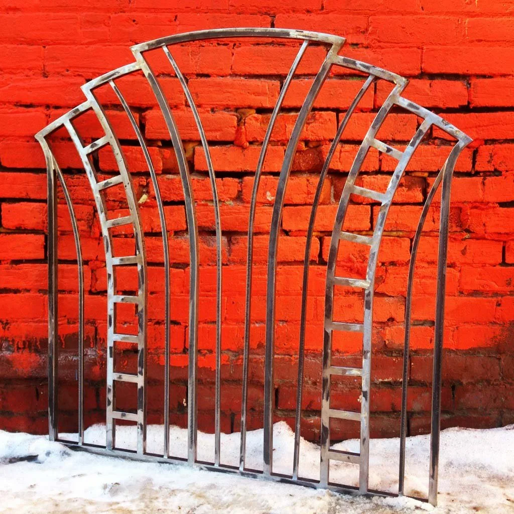 A metal gate with an arched top and ladder-like bars, leaning against a red brick wall with snow on the ground.