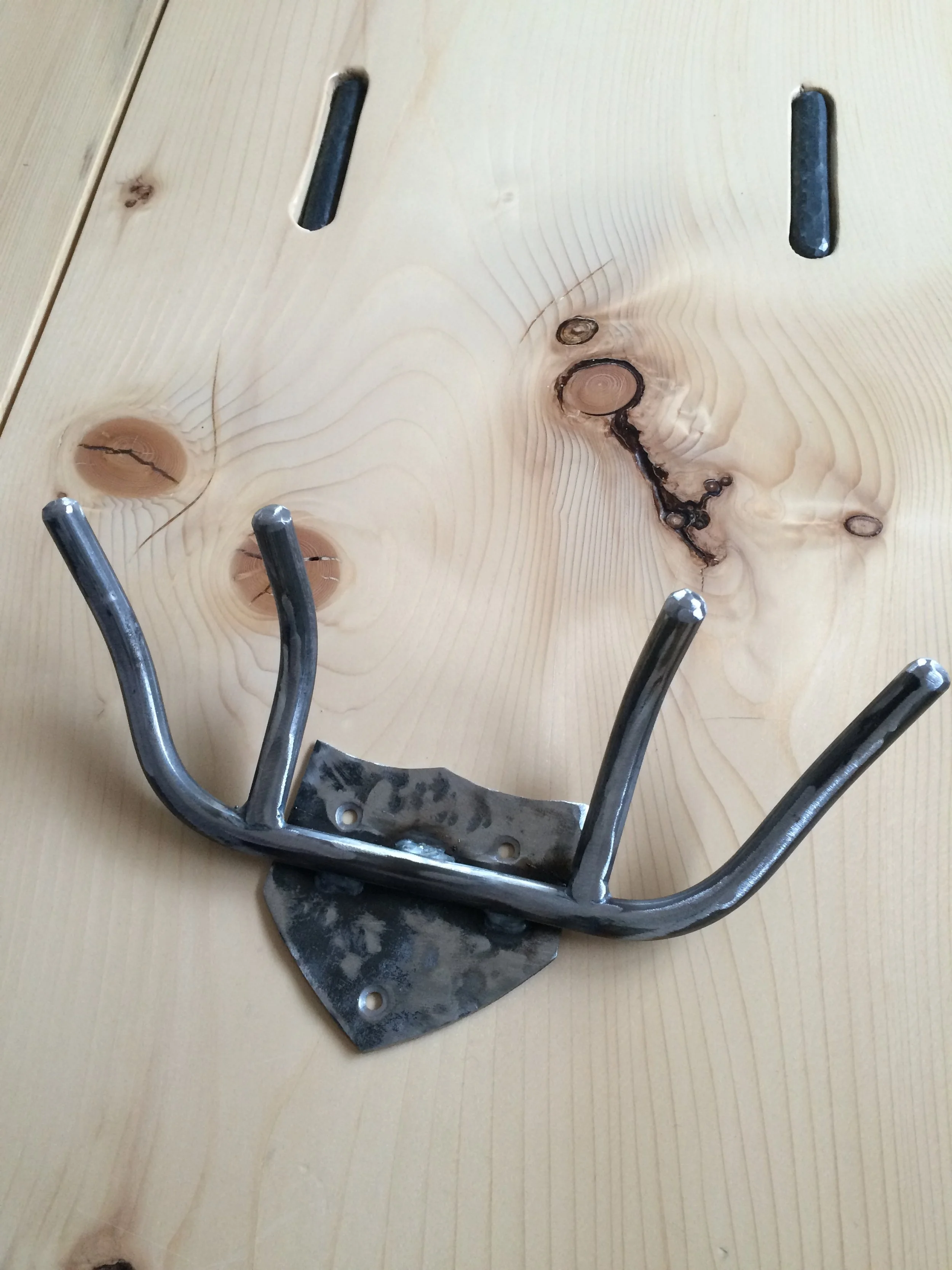 A metal wall hook with four curved arms attached to a metal plate, placed on a light-colored wooden surface with visible wood grain and knots.