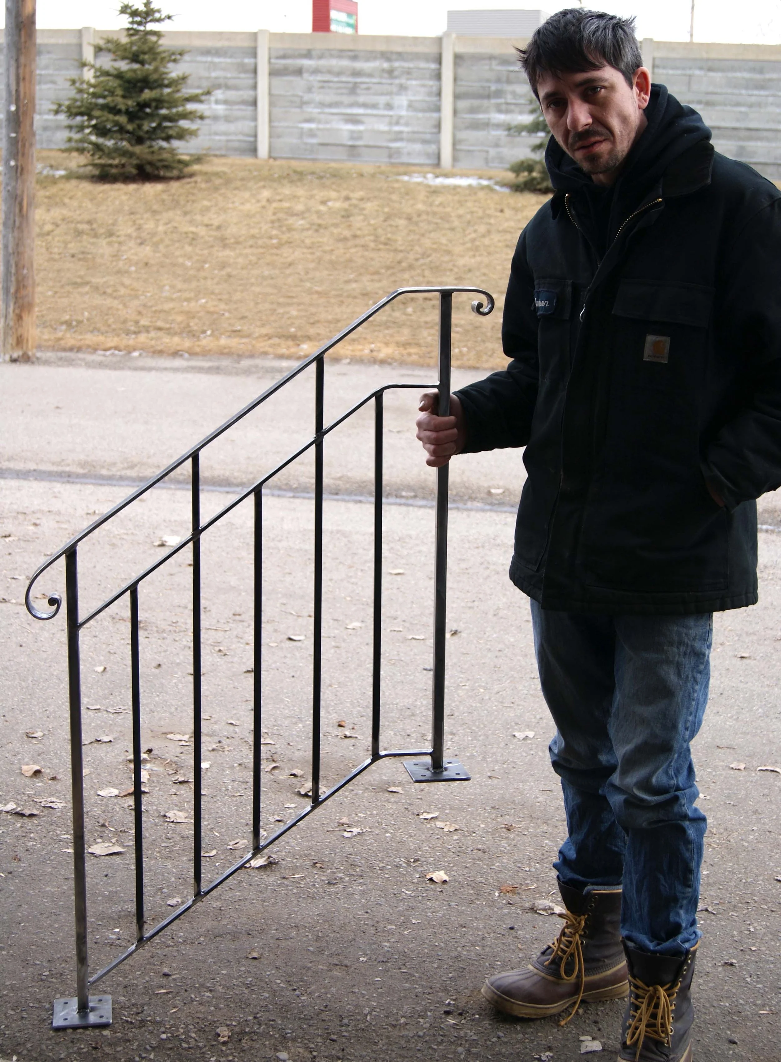 A man standing outdoors on a sidewalk holding a metal gate. He wears a black jacket, jeans, and brown boots, with a neutral expression on his face.