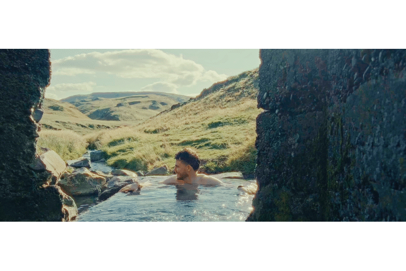 A man enjoying a hot spring in a scenic outdoor landscape with rolling hills and a partly cloudy sky.