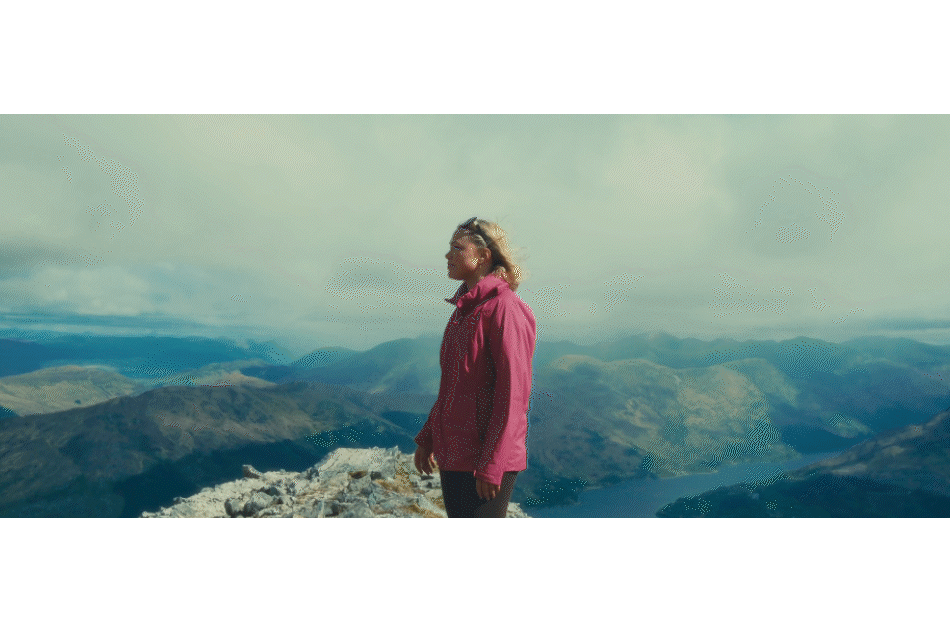 A woman in a pink jacket standing on a mountain peak overlooking a scenic landscape of mountains and clouds.