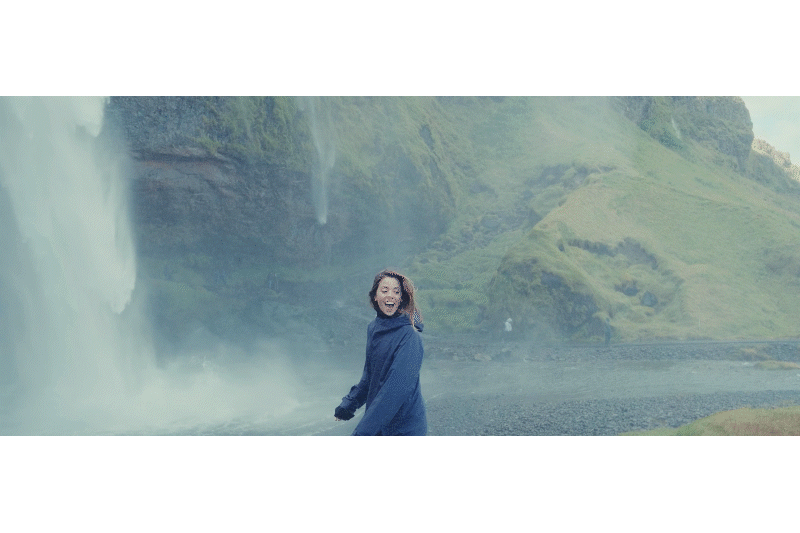 A woman wearing a blue jacket standing near a large waterfall with lush green landscape in the background.