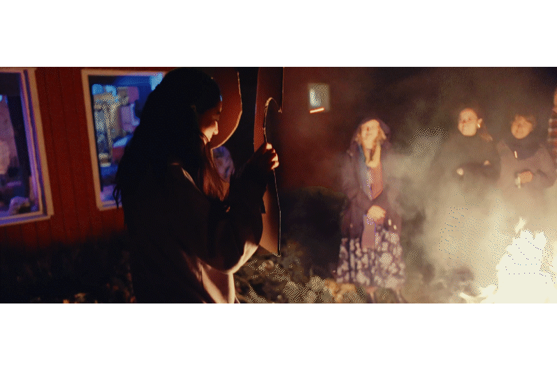 Person holding a bow and arrow at a nighttime outdoor gathering with a fire and several onlookers