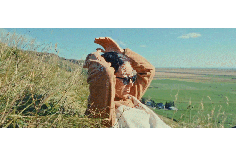 Woman lying on grassy hill, wearing sunglasses and a tan jacket, with wide open landscape and blue sky in the background.