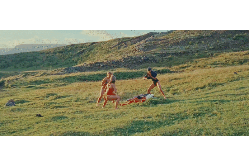 Three women on a grassy hillside, with one woman kneeling and two standing, engaging in a playful or competitive activity. The background features rolling hills and a partly cloudy sky.