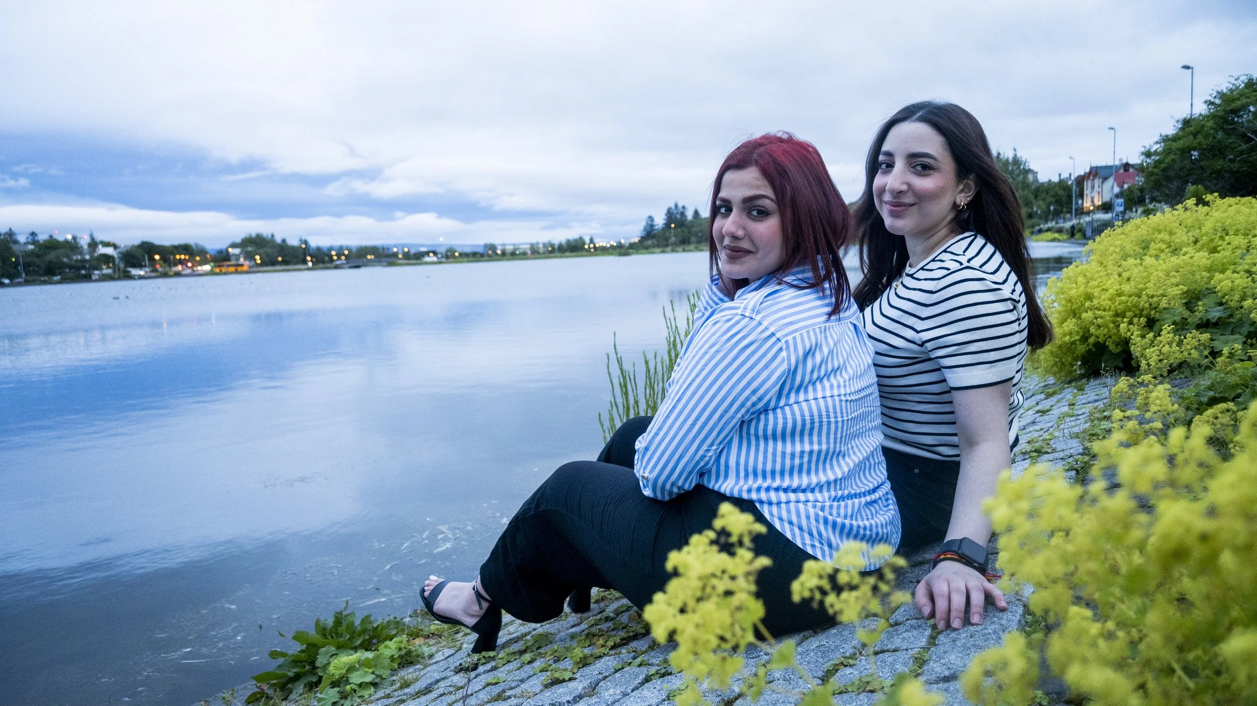Two women sitting by a lakeside at dusk, with buildings and trees in the background, and yellow-green plants in the foreground.