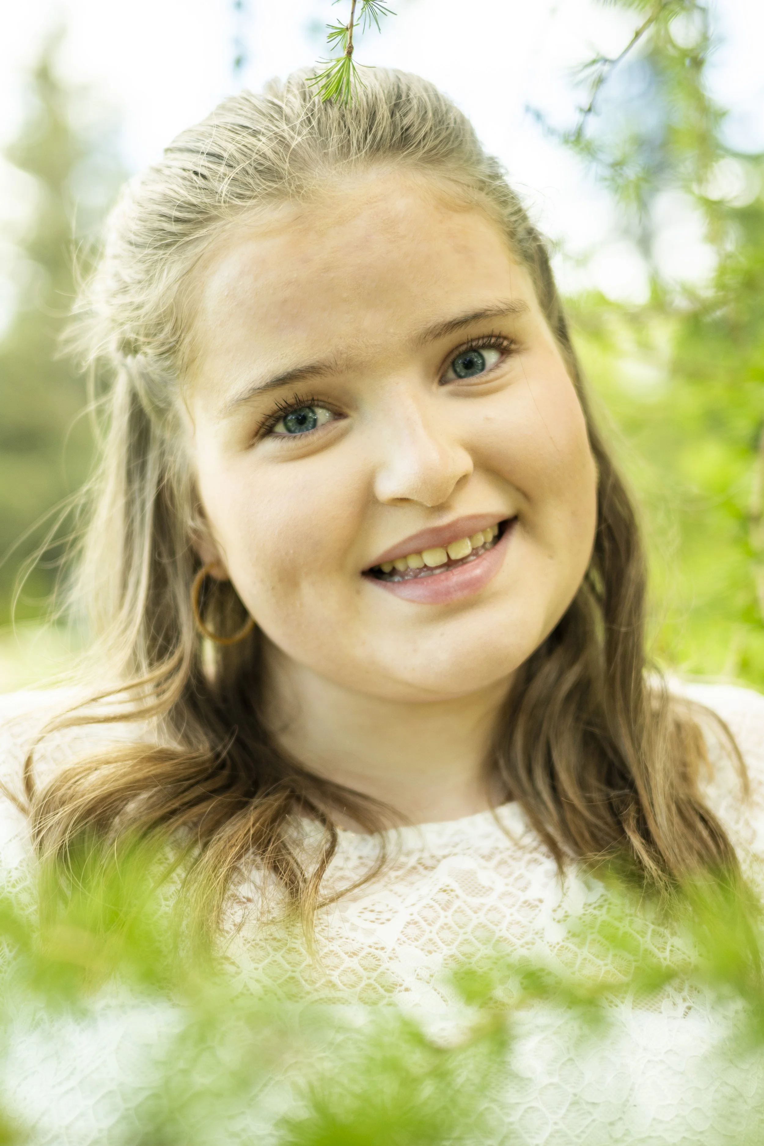 Close-up of a young woman with long curly hair and blue eyes, smiling outdoors among green foliage and trees.