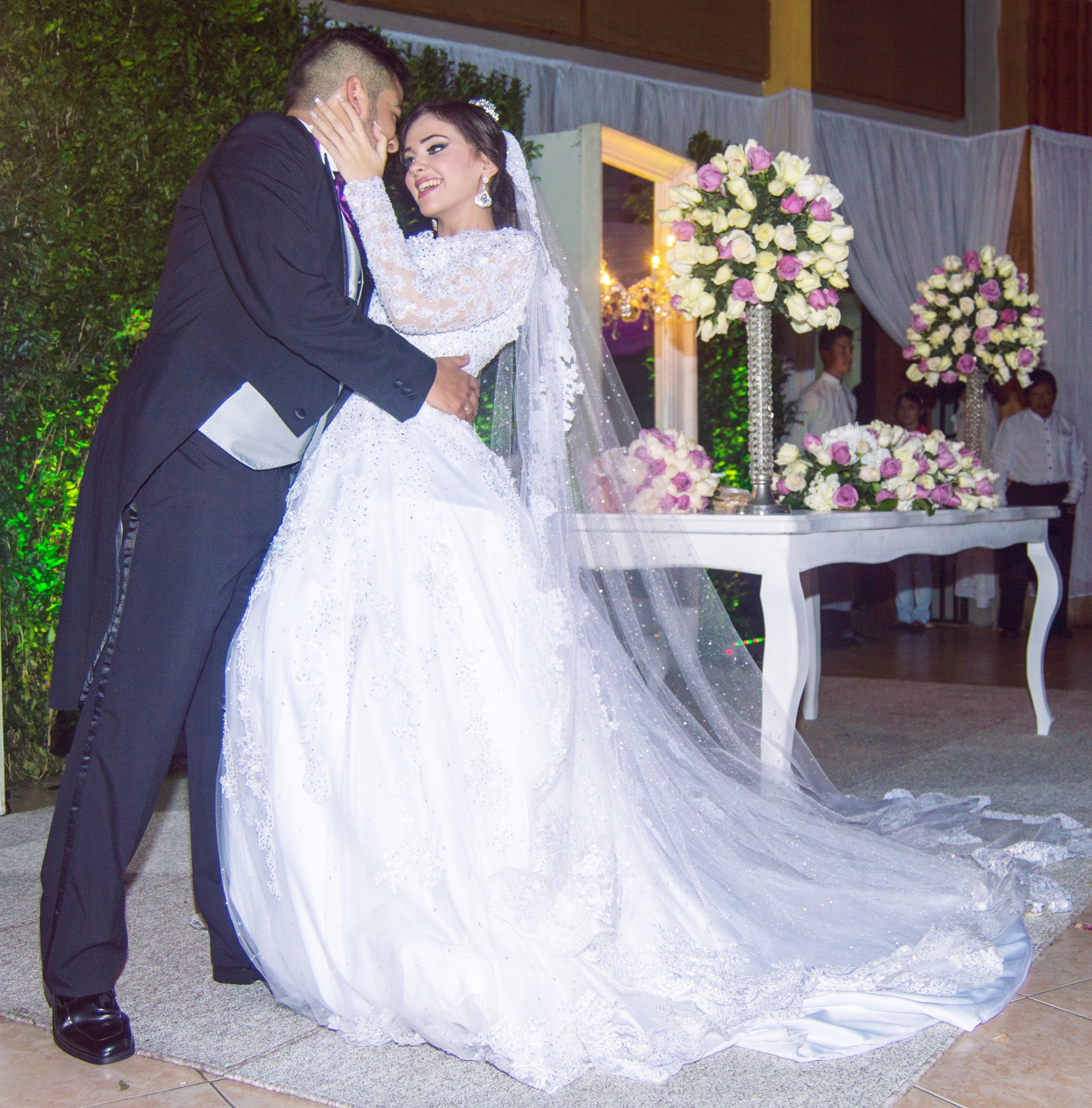 A bride and groom sharing a romantic moment at their wedding reception. The bride is wearing a white gown and veil, and the groom is in a black tuxedo. They are standing near a table decorated with pink and white floral arrangements.