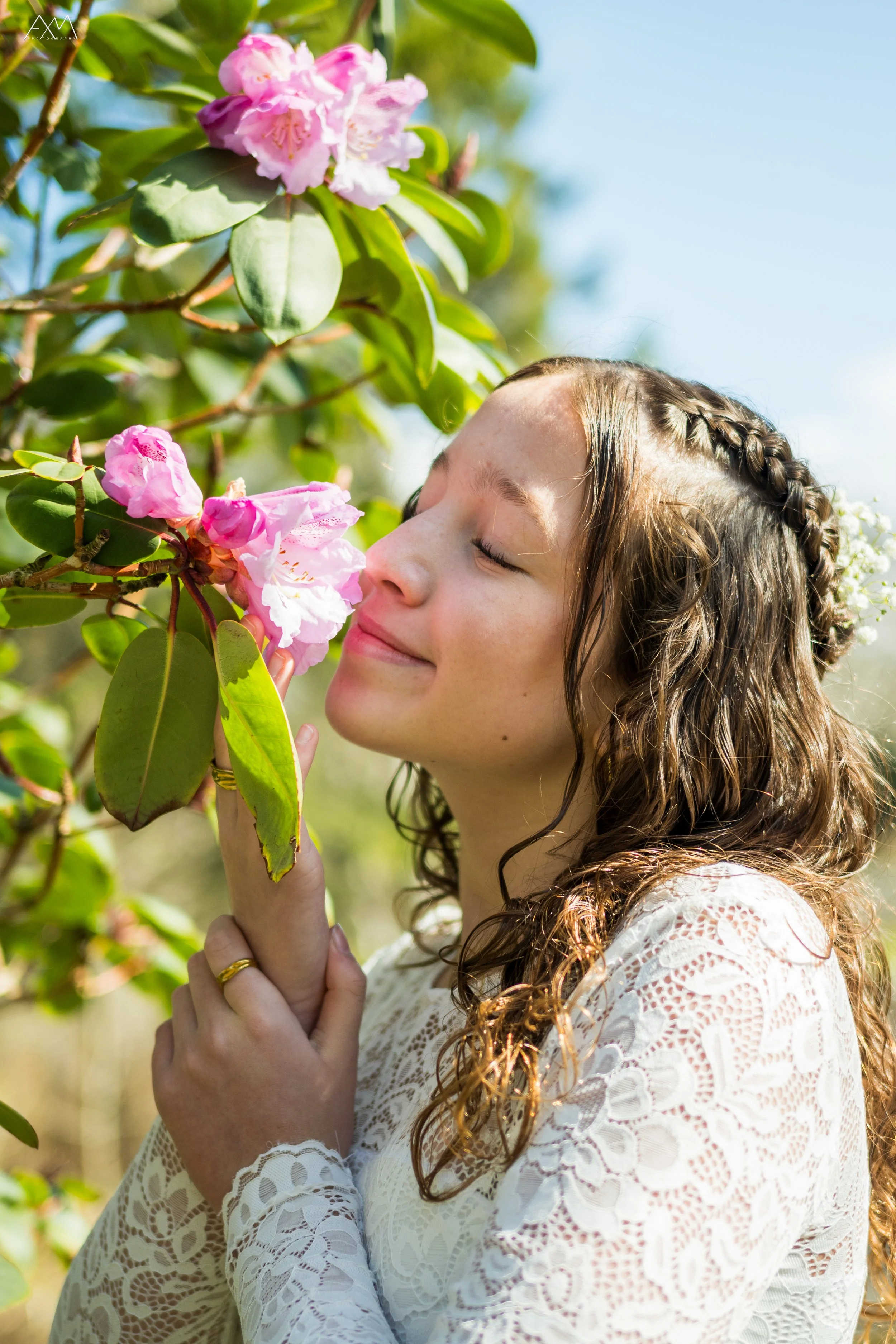 A woman with curly brown hair and closed eyes is smiling and gently smelling pink flowers on a green bush outdoors, wearing a white lace dress.