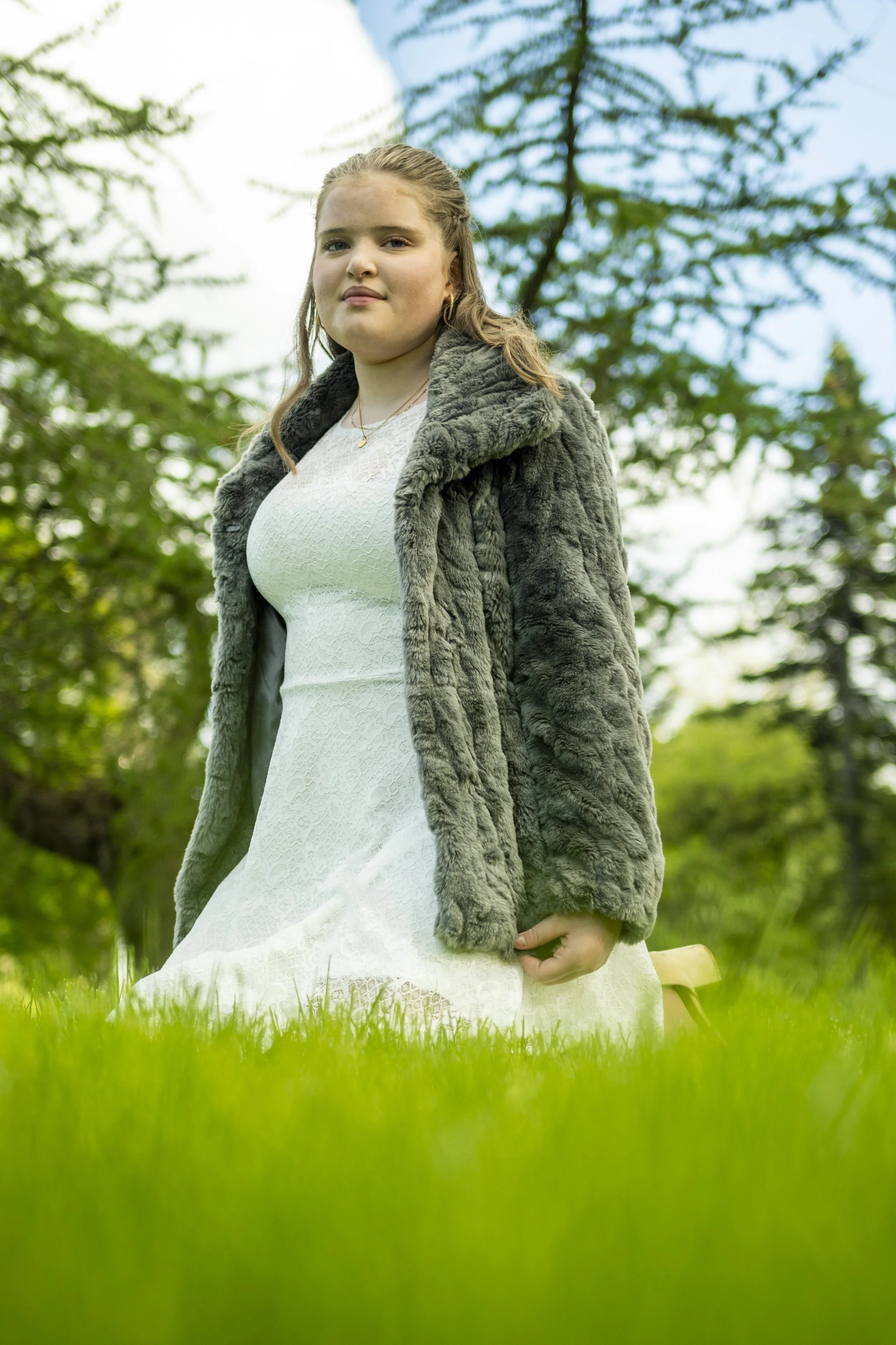 A young woman with light brown hair in a braid, wearing a white lace dress and a gray faux fur coat, sitting outdoors on grass with blurry green trees and a blue sky in the background.
