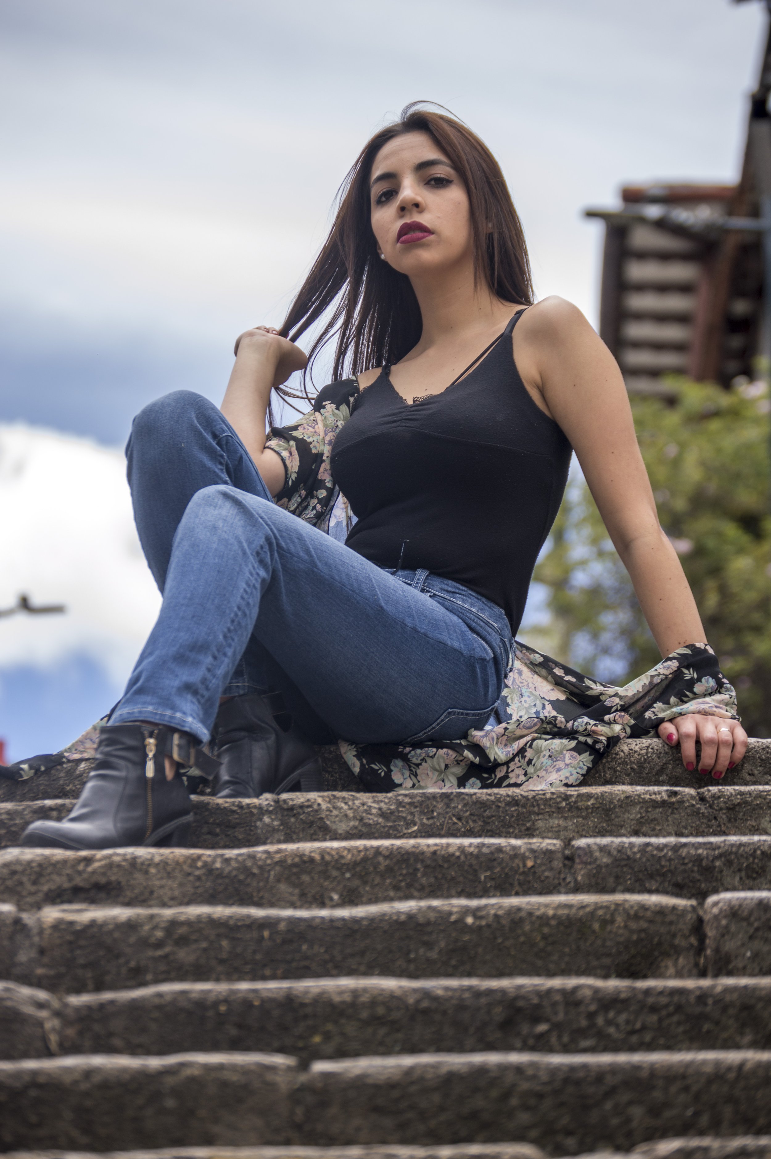 A young woman with long brown hair and fair skin sitting on stone steps outdoors, wearing a black tank top, blue jeans, and black ankle boots, with a floral scarf draped over her shoulder. She is looking at the camera with a neutral expression, again