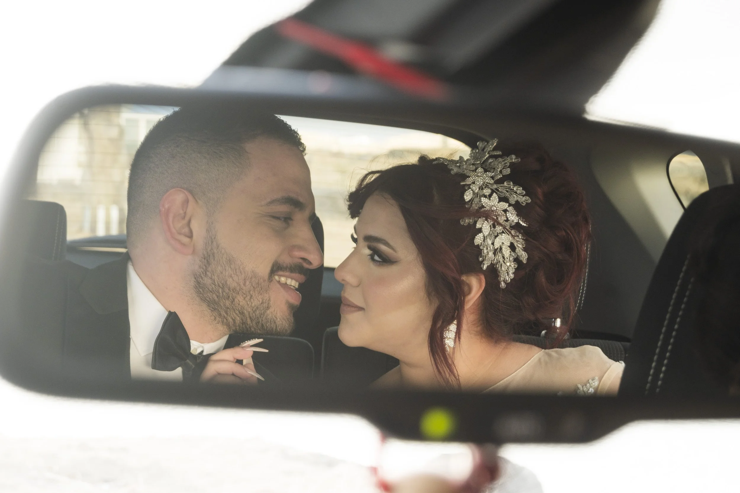 A bride and groom sitting close together in the backseat of a car, looking into each other's eyes with smiles, the bride wearing a decorative hairpiece.