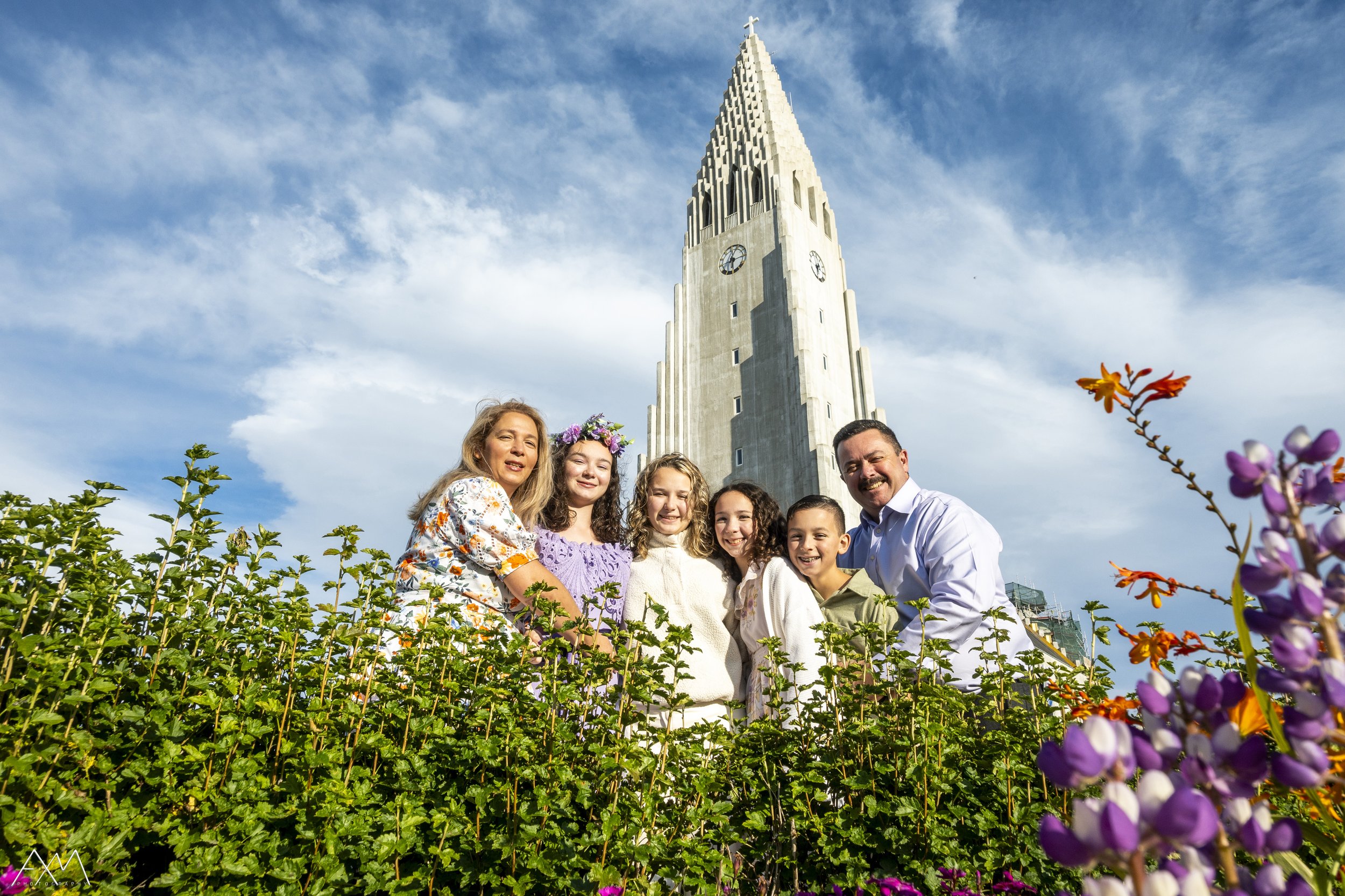 A family of six standing behind green bushes with purple flowers in front of Hallgrímskirkja church tower in Reykjavik, Iceland, on a partly cloudy day.