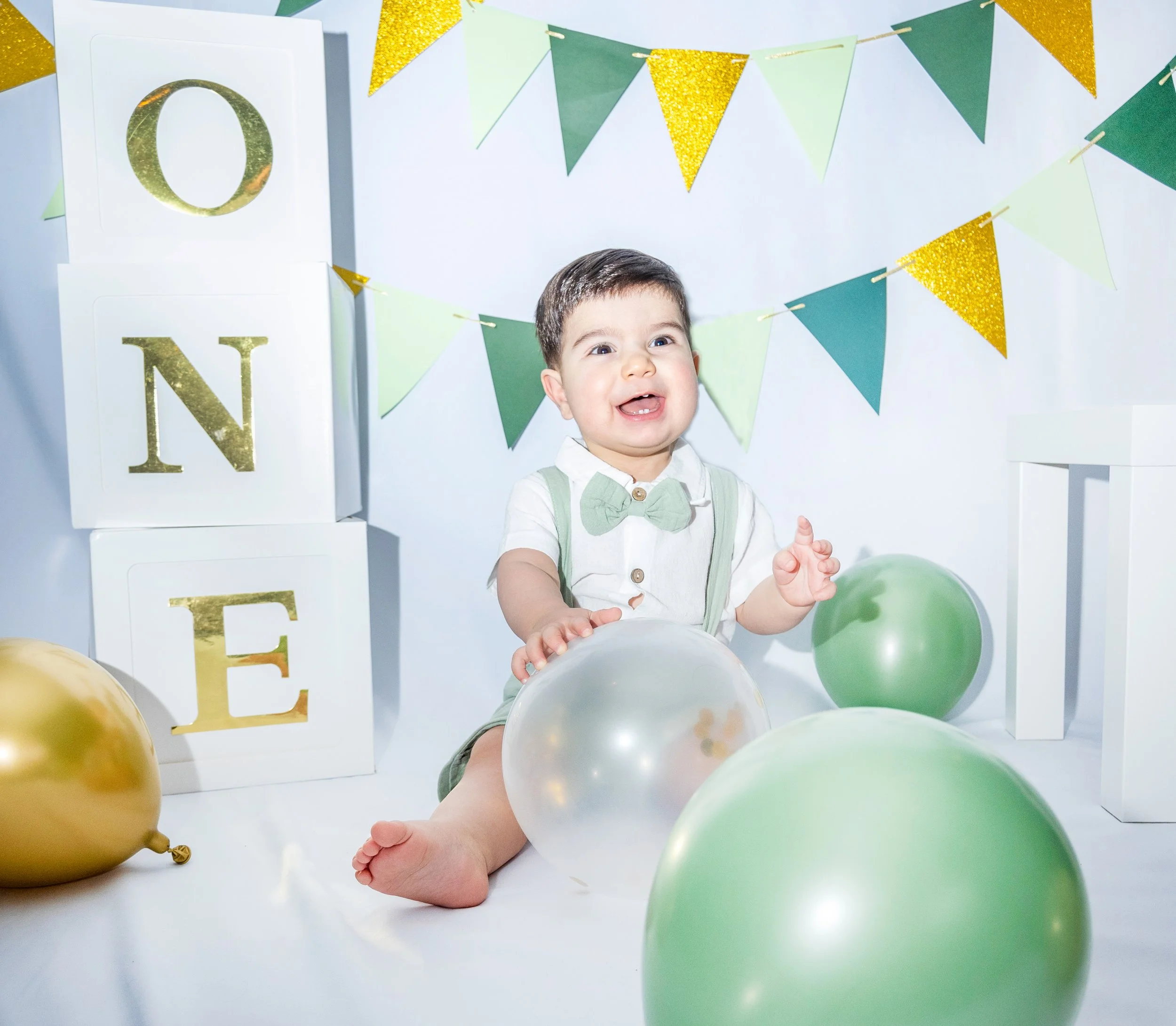 Child smiling at birthday celebration with green and gold balloons, gold and green bunting, and white blocks with gold and green letters.