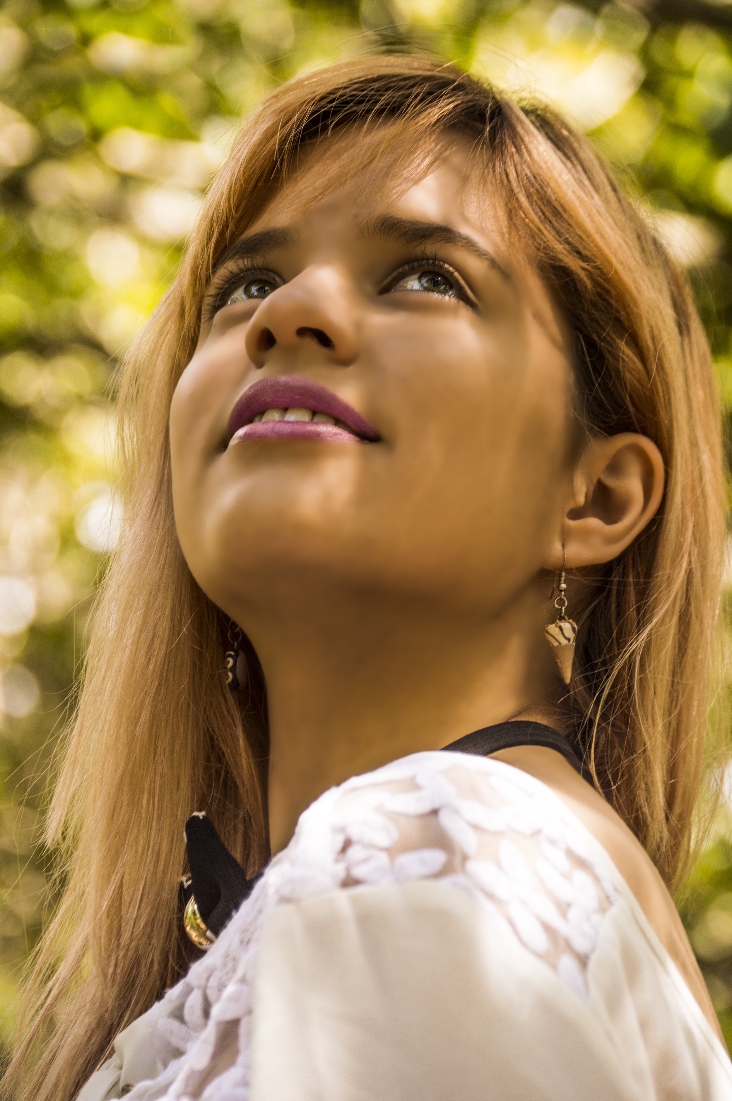 Close-up of a young woman with long reddish hair, wearing earrings and a white top, looking upwards outdoors during daytime with sunlight filtering through green leaves in the background.