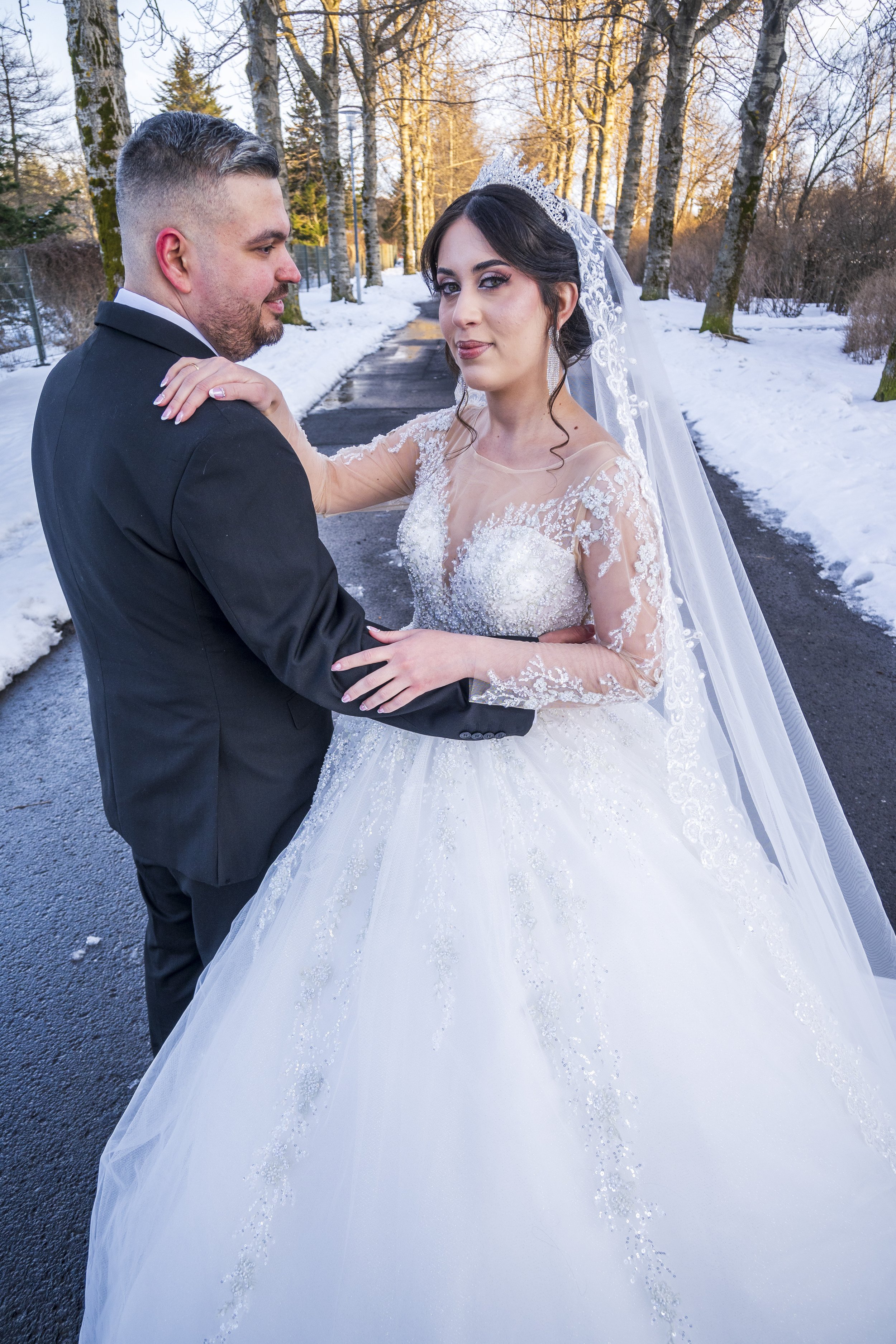 A bride and groom standing on a snowy pathway outdoors with trees in the background, dressed in wedding attire, during sunset.