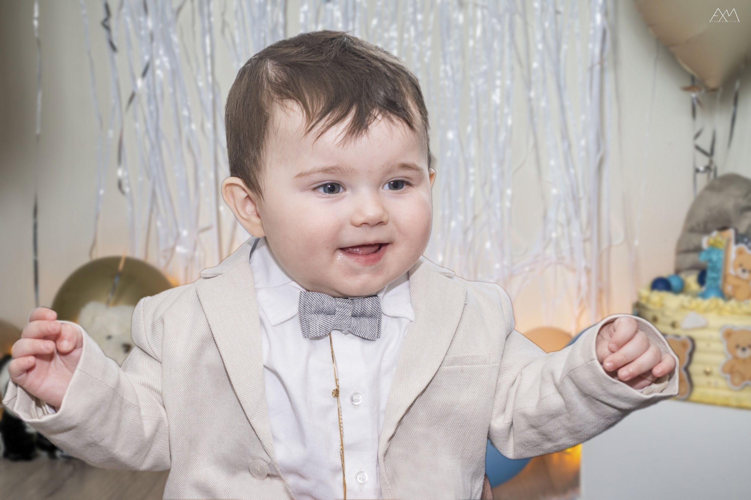 A young boy with light brown hair and blue eyes, wearing a light beige blazer, white shirt, and gray bow tie, celebrates at a birthday party with a decorated background of balloons and metallic streamers.