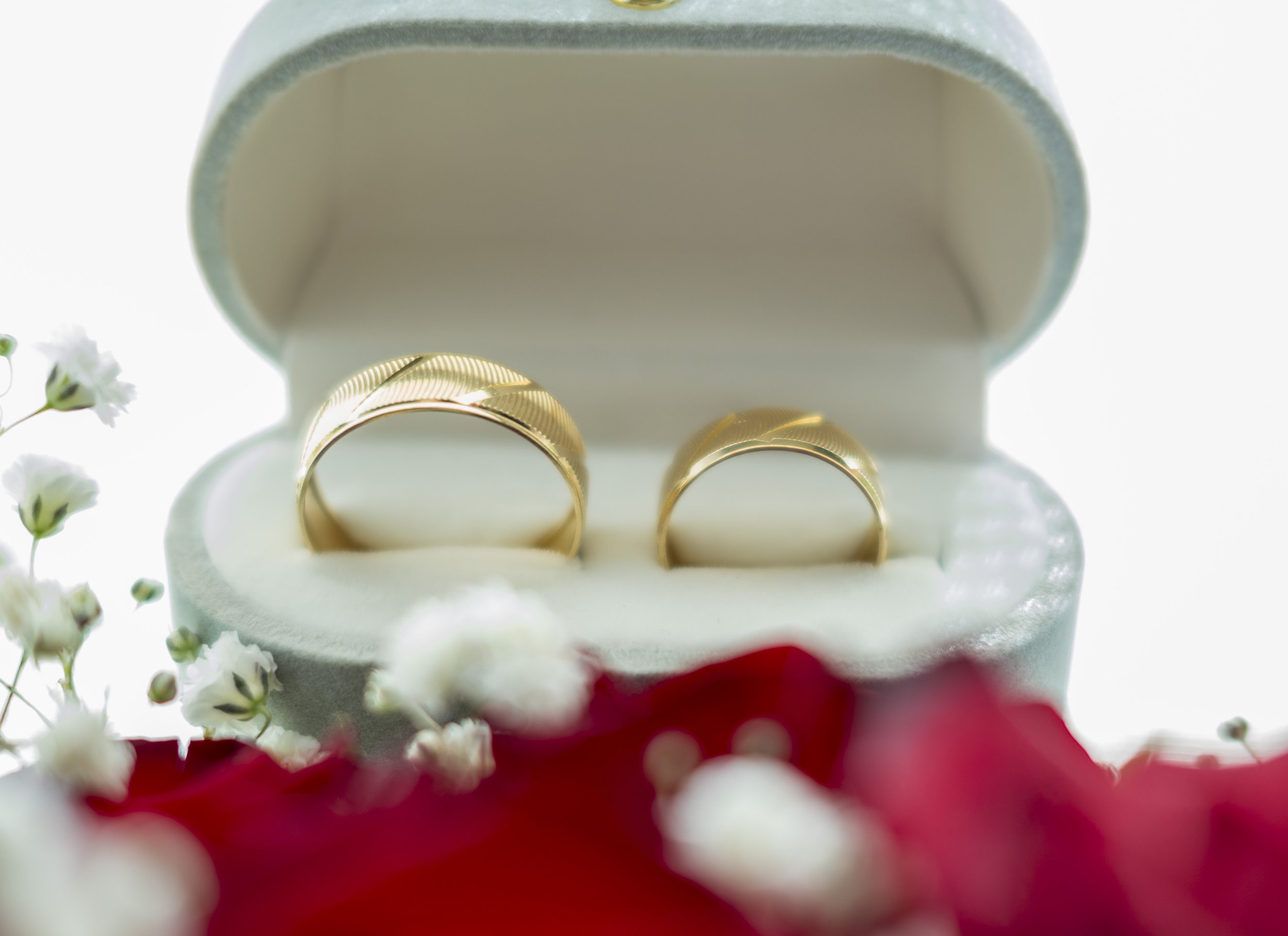 Gold wedding rings in an open white jewelry box, surrounded by white and pink flowers.