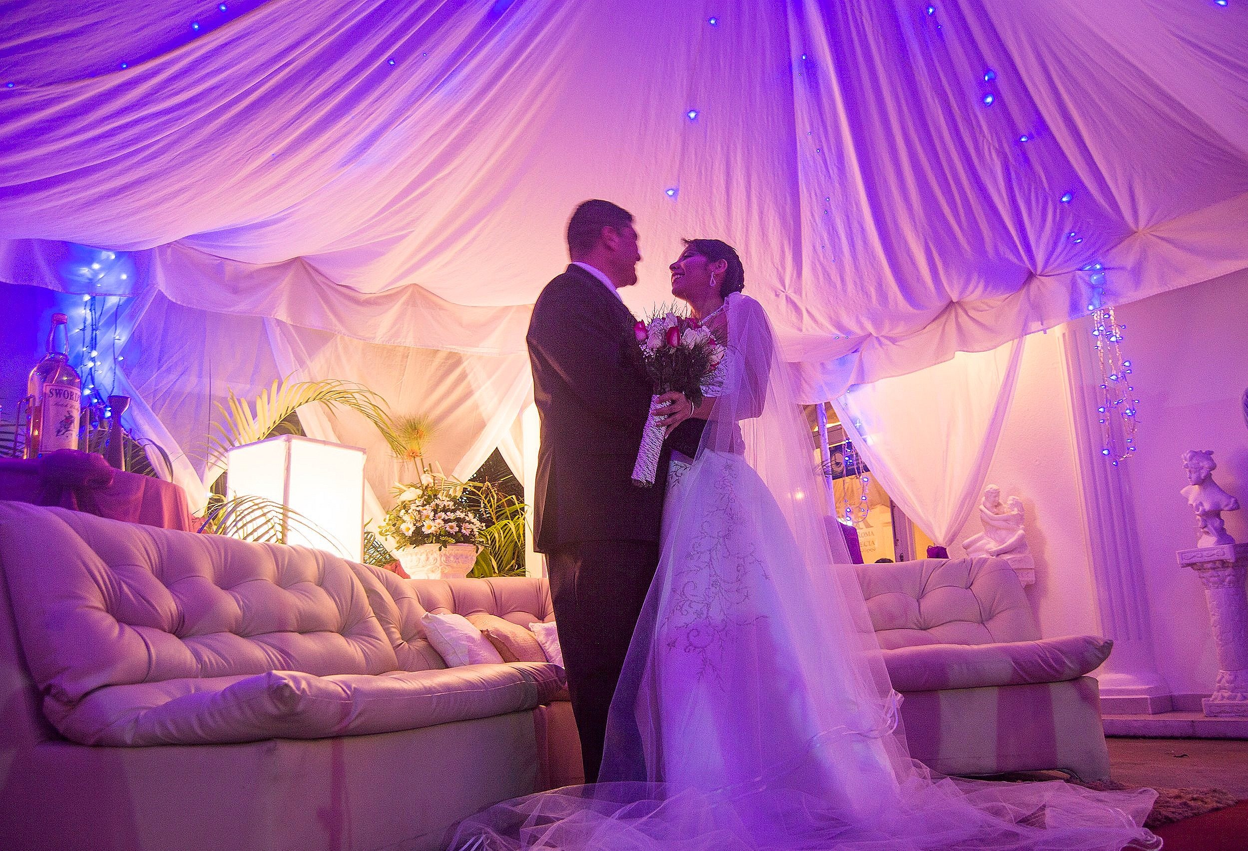 A bride and groom standing under a decorated canopy, smiling at each other, with the bride holding a bouquet of flowers, during a wedding celebration.