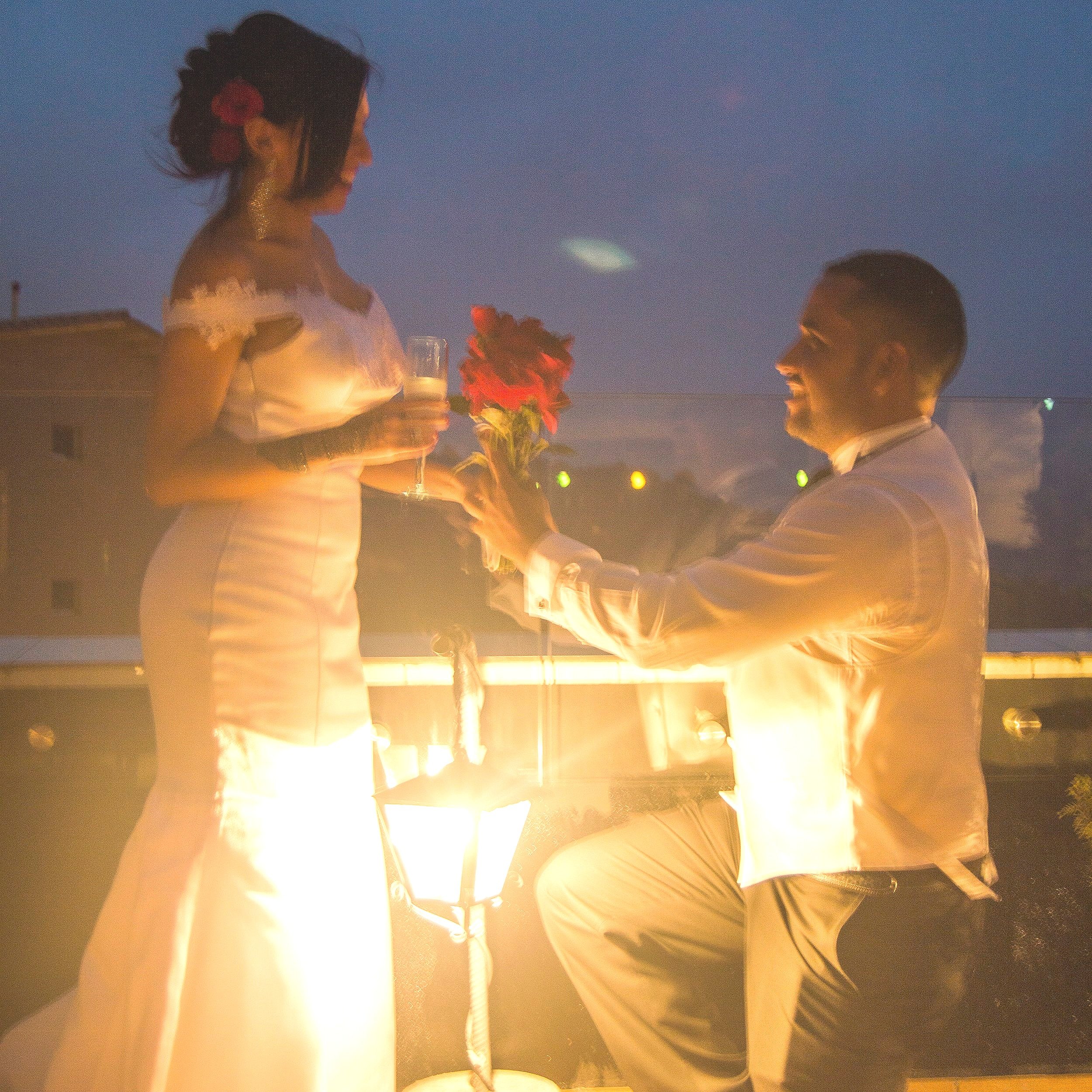 A man is proposing to a woman on a balcony during the evening. He is kneeling and holding a bouquet of red flowers, while she is holding a glass of champagne. The scene is lit by a street lamp and has a romantic ambiance.
