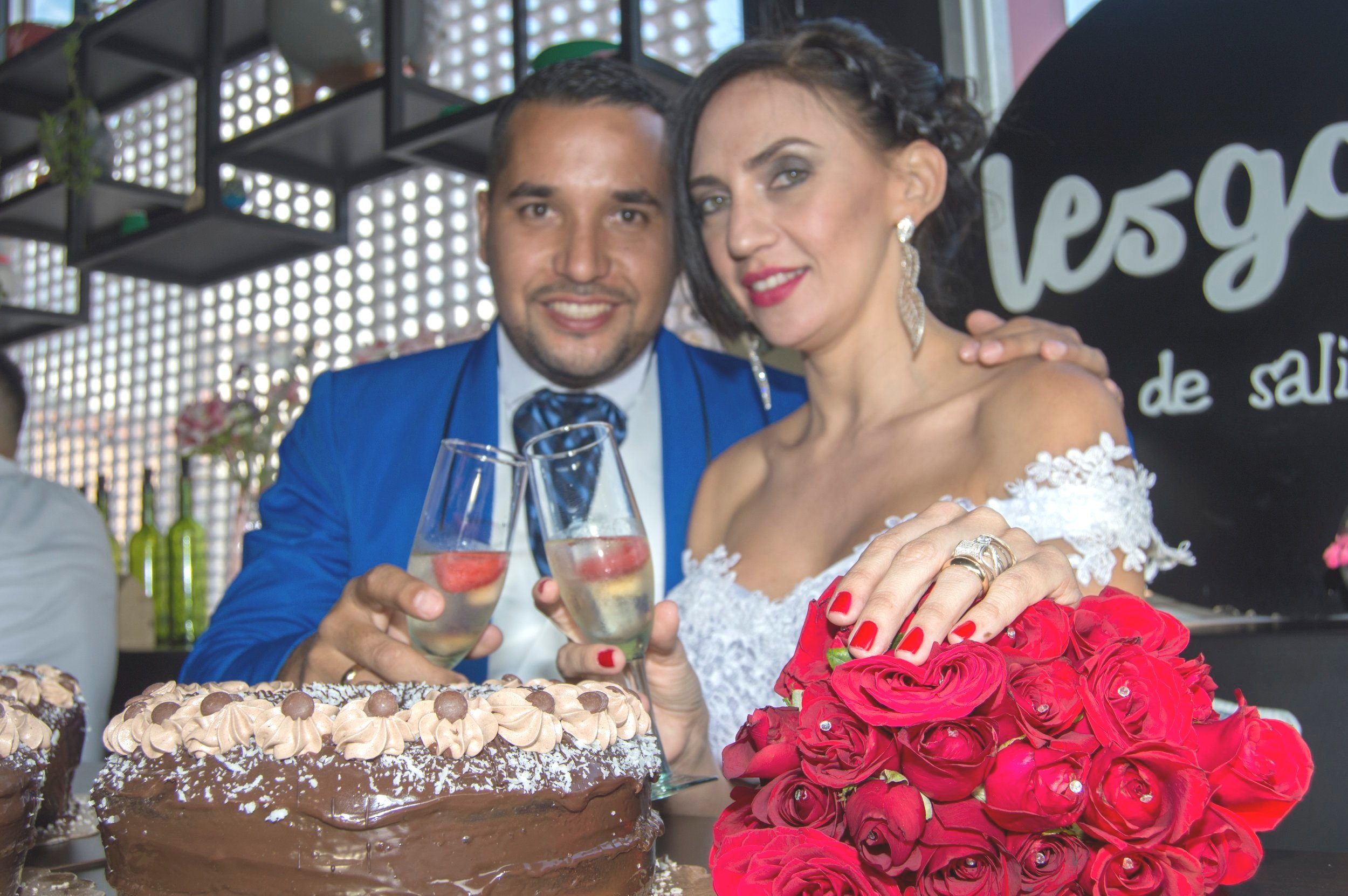 A newlywed couple at their wedding celebration, holding champagne glasses, with wedding cake and a bouquet of red roses in front of them.