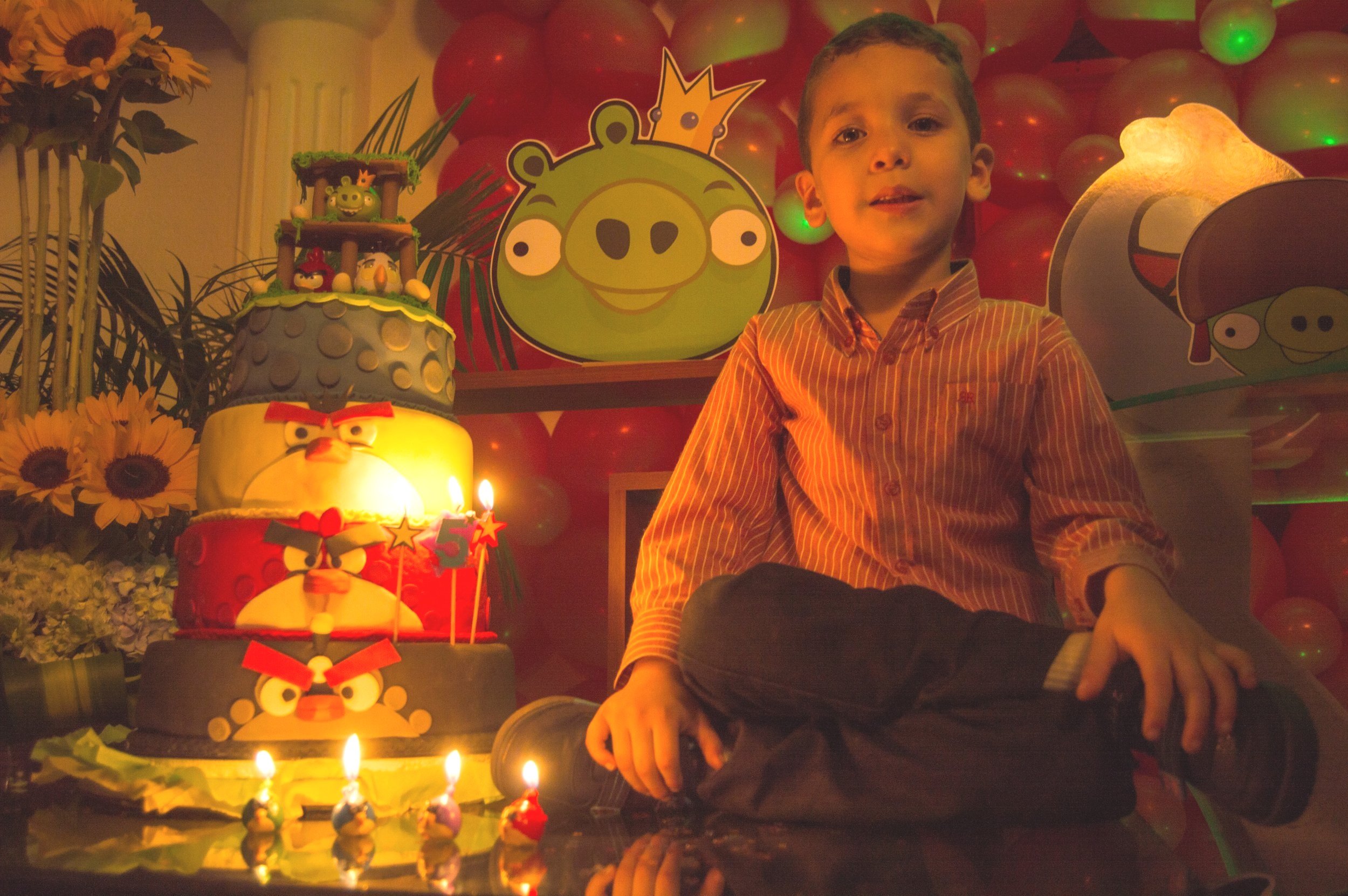 A young boy sitting cross-legged in front of a multi-layered Angry Birds themed birthday cake with lit candles. The background features colorful balloons, an Angry Birds character face cutout, and strategic Angry Birds decorations, including a large