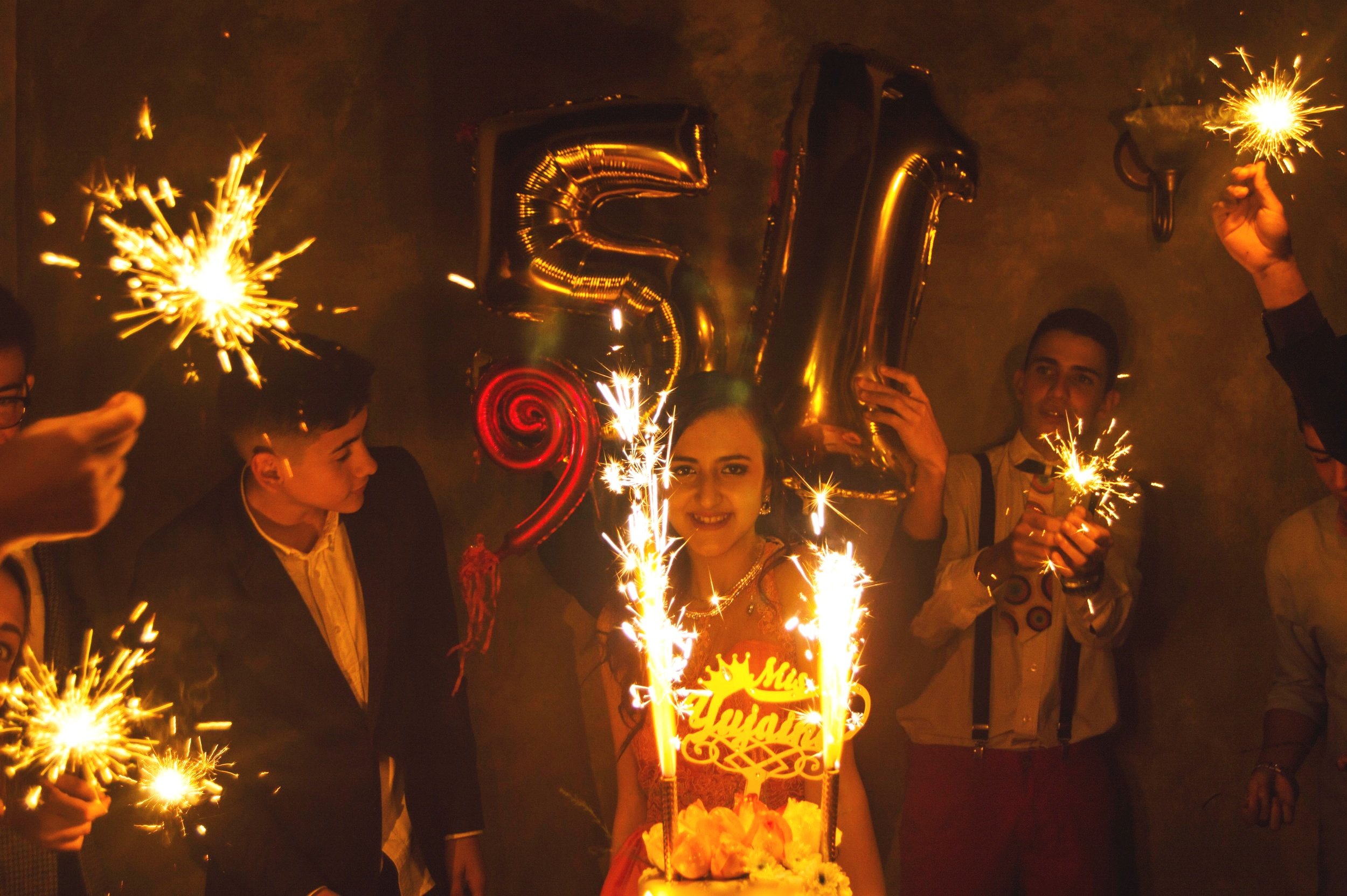 A woman celebrating her 25th birthday with friends, holding balloons that spell out 25, surrounded by sparklers, and a birthday cake with candles, in a festive indoor setting.