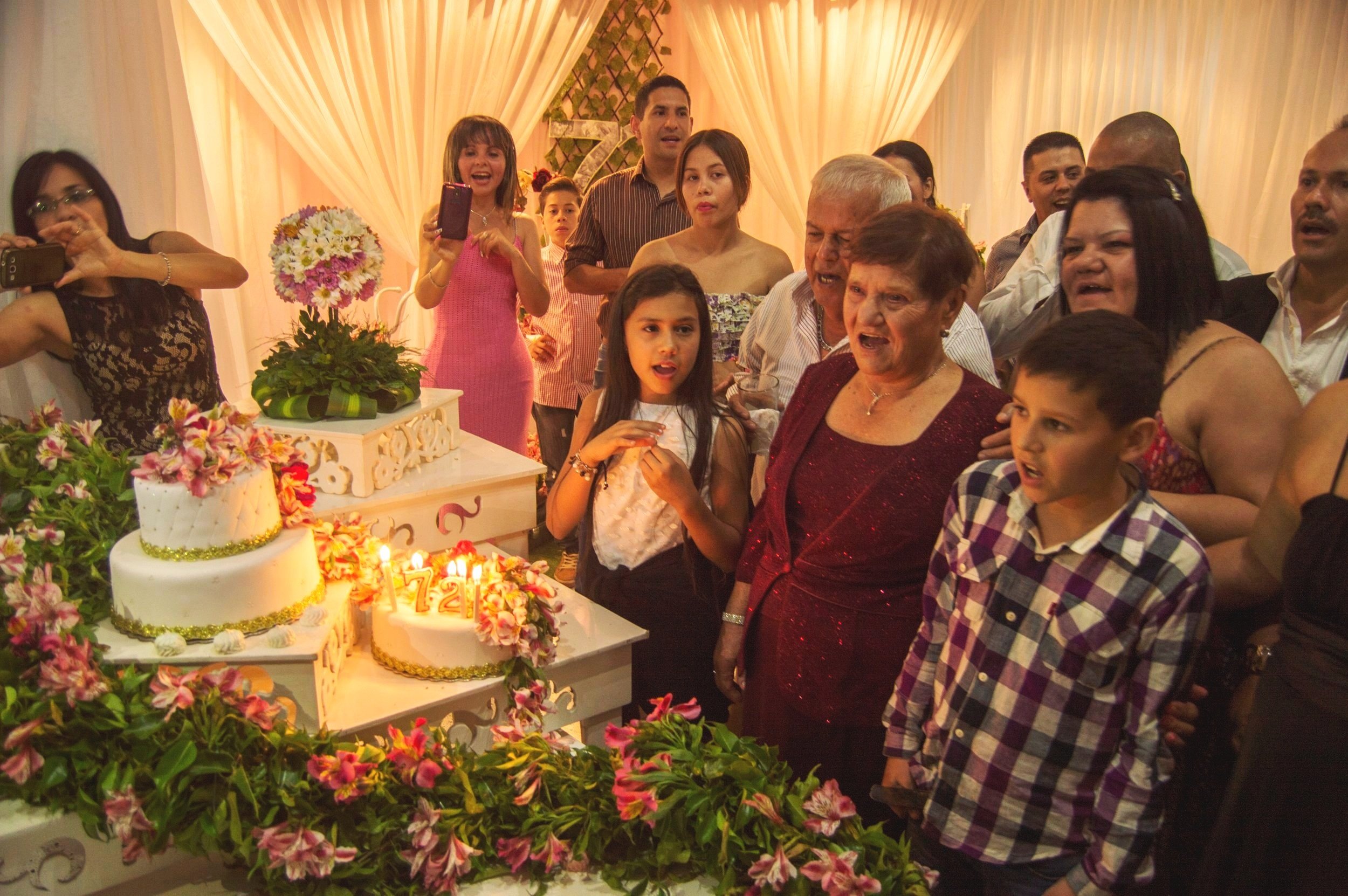 A family celebrating a birthday with a decorated cake and lit candles, surrounded by guests taking photos and enjoying the party indoors with floral decorations.