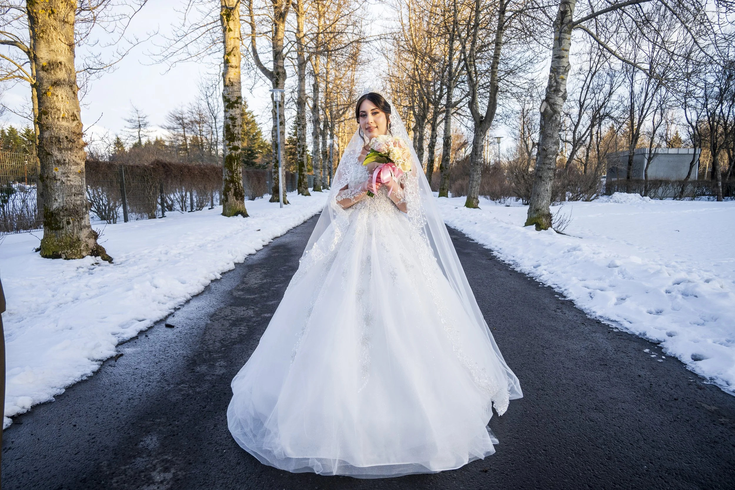 A bride in a white wedding dress holding a bouquet of flowers standing on a snow-covered path surrounded by leafless trees during winter.