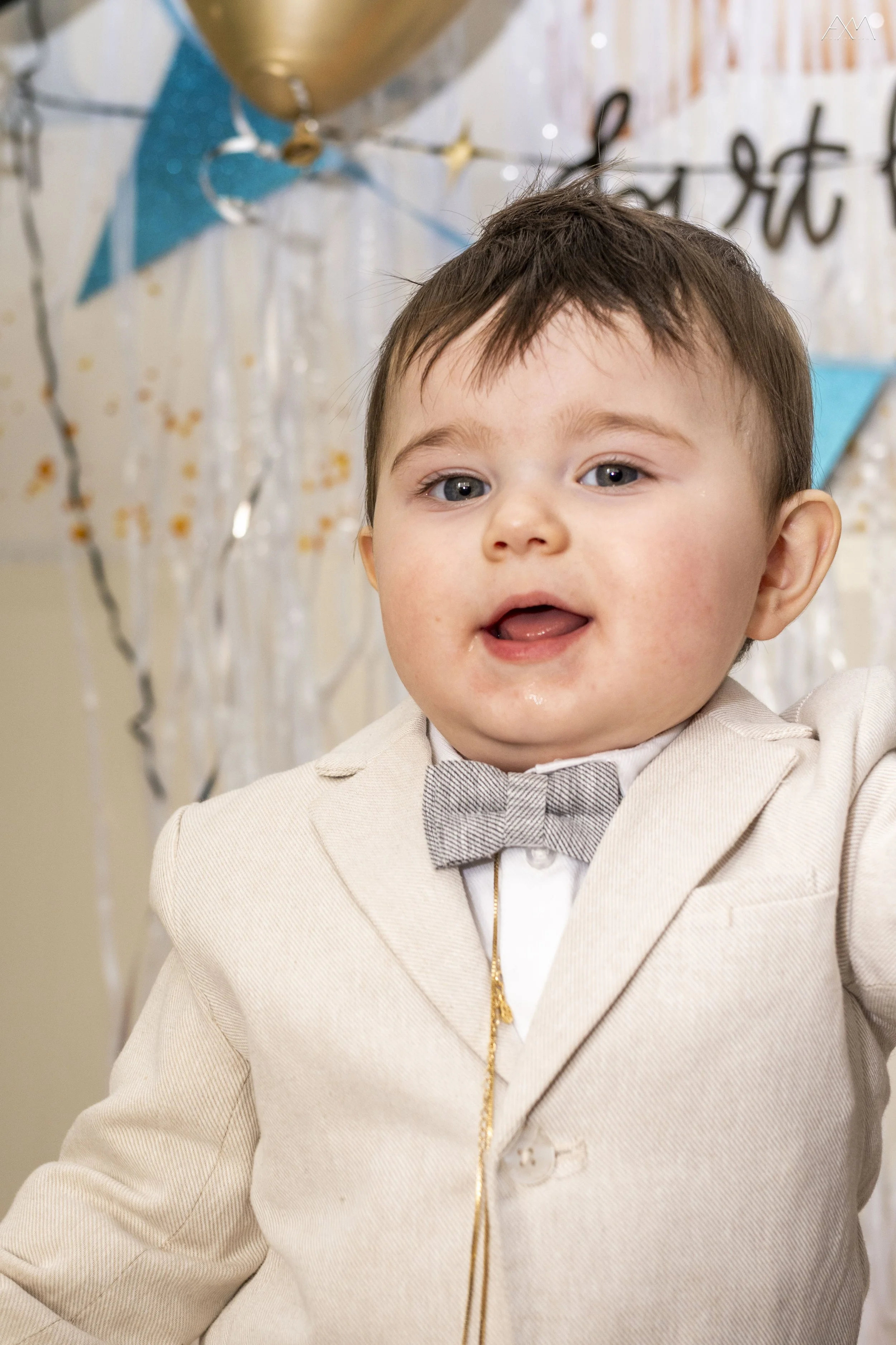 A young boy with brown hair and blue eyes, wearing a beige tuxedo with a bow tie, at a celebration event decorated with balloons and banners.