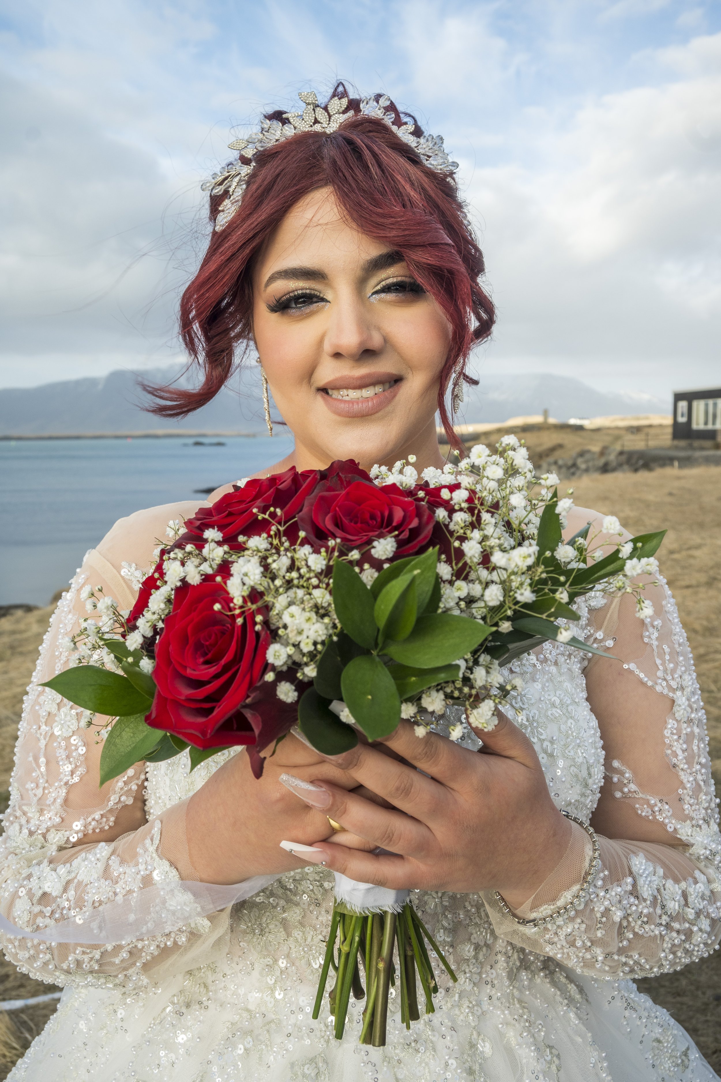 A woman in a wedding dress holding a bouquet of red roses and white flowers by a body of water with mountains in the background.