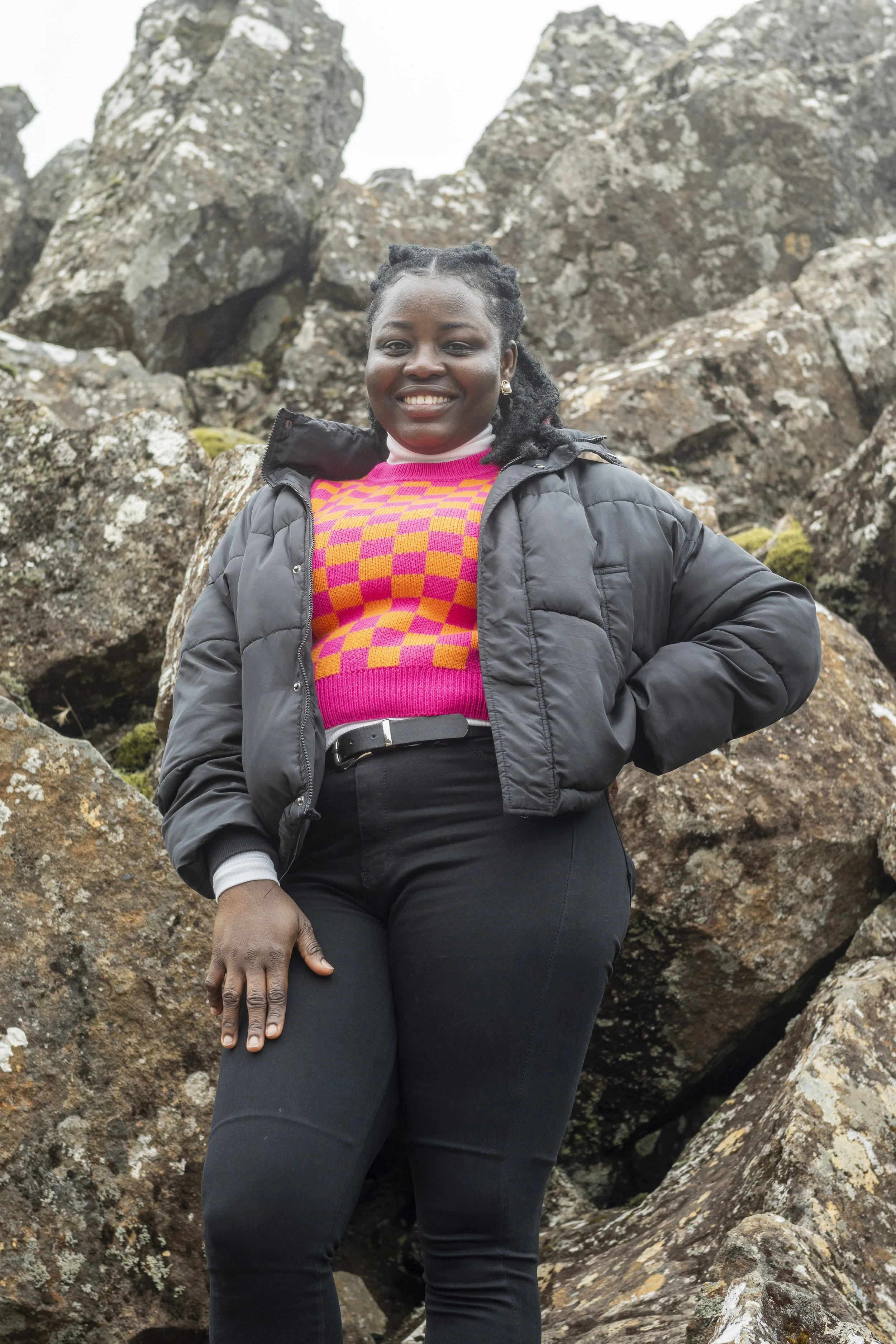 A woman standing outdoors among large rocks, smiling at the camera, wearing a black puffer jacket, a colorful pink and orange checkered sweater, and black pants.