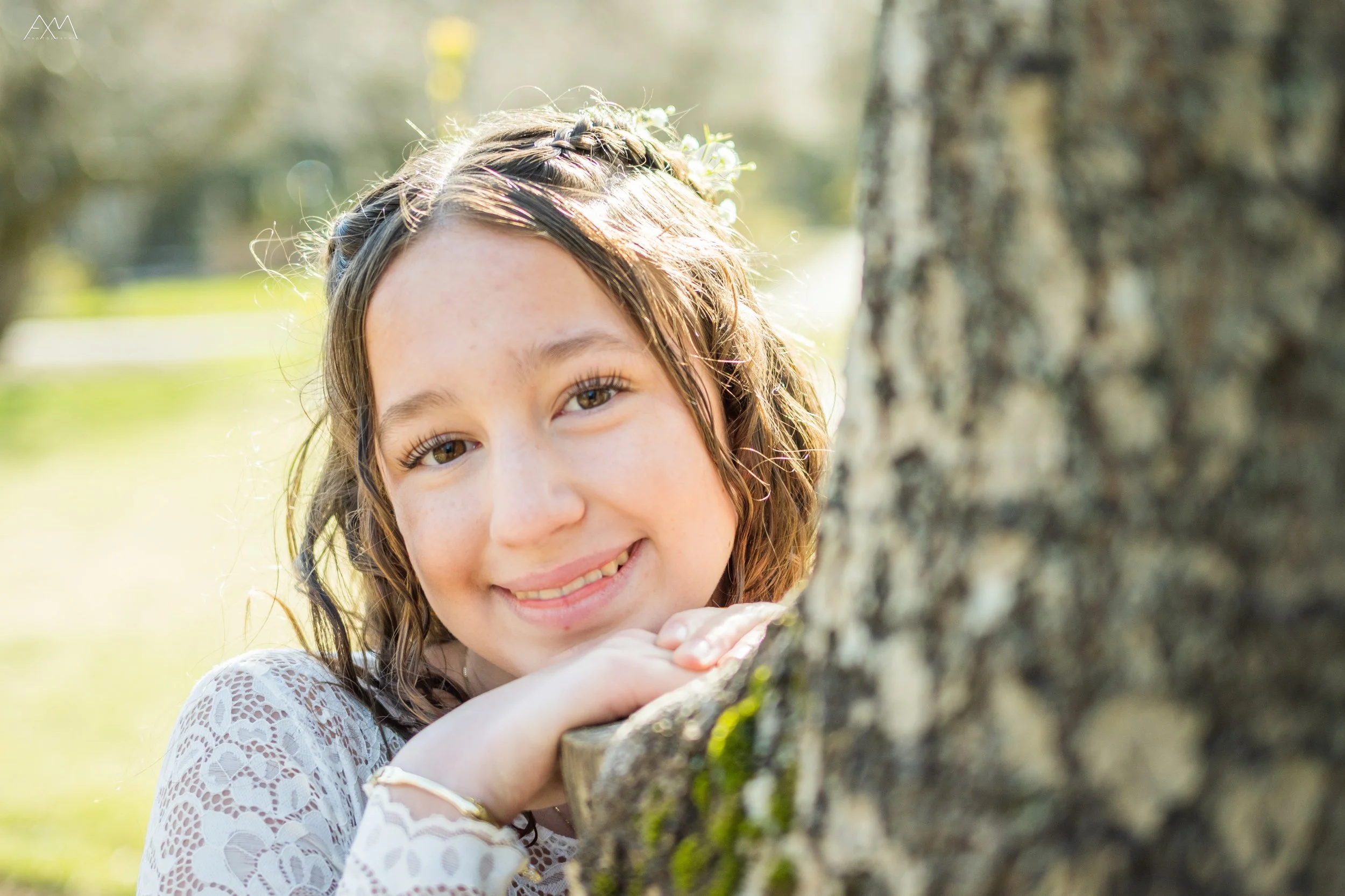 A young girl with brown curly hair smiling behind a tree trunk outdoors.