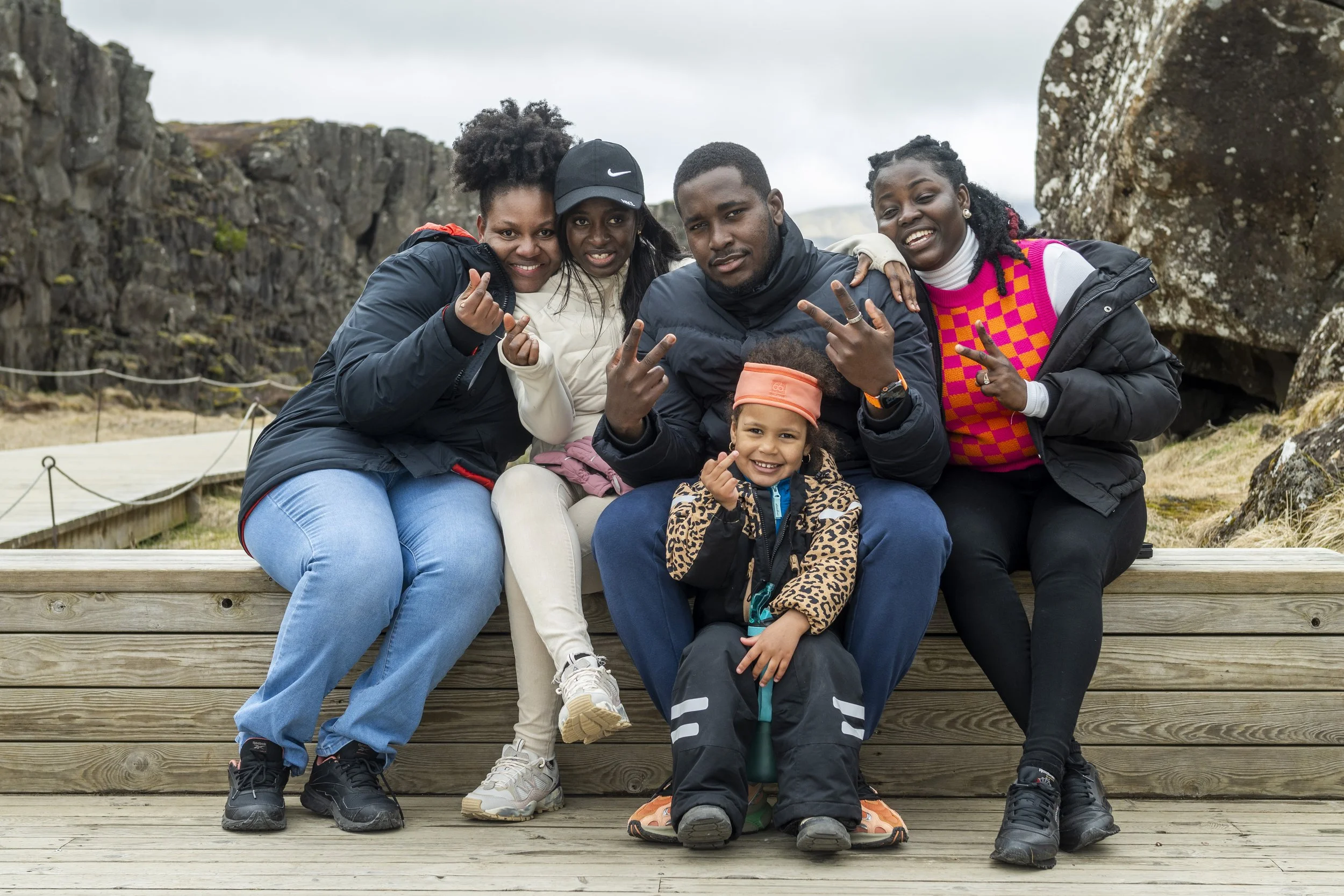 Group of five friends sitting on a wooden bench outdoors, smiling and making hand gestures, with rocky landscape and overcast sky in the background.