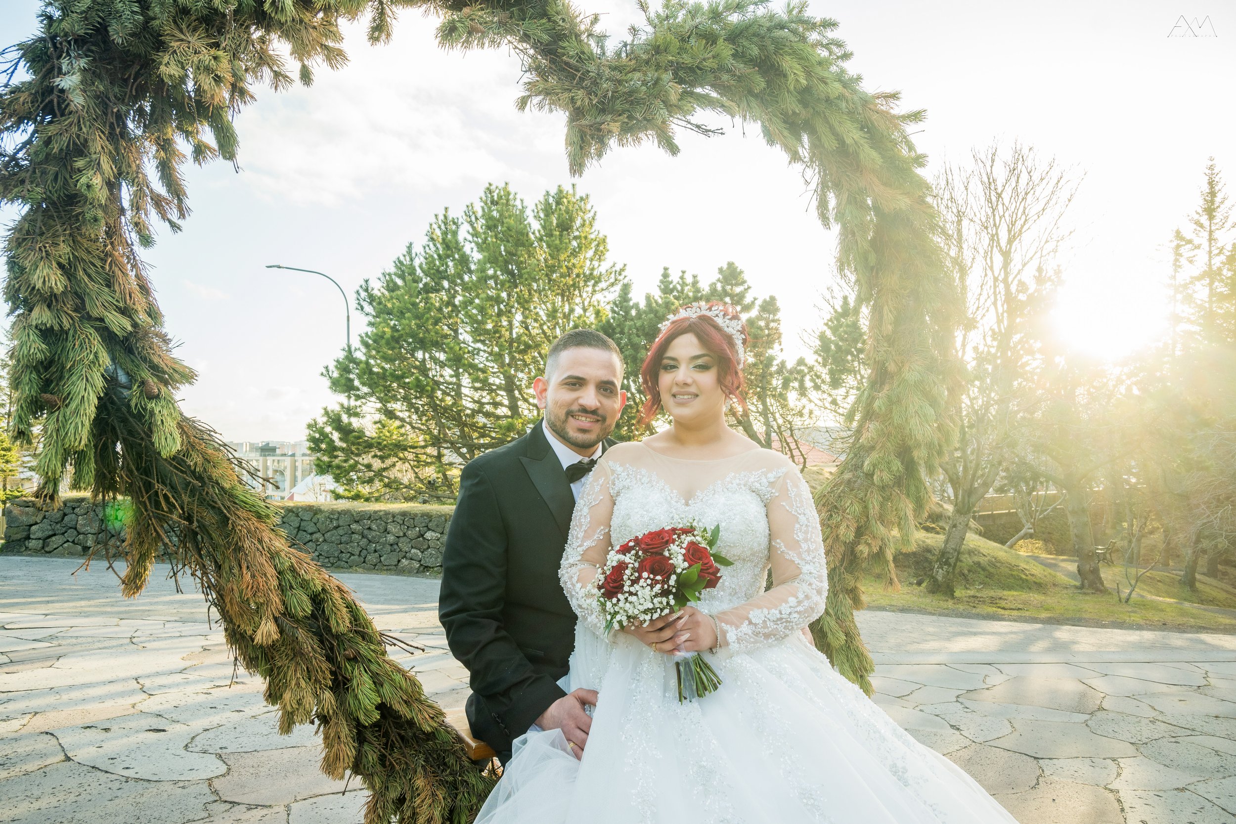 A bride and groom sit on a bench beneath a large, decorated arch in a park during their wedding photos. The bride has red hair and is wearing a white wedding gown holding a red rose bouquet. The groom is dressed in a black tuxedo. The sun is shining