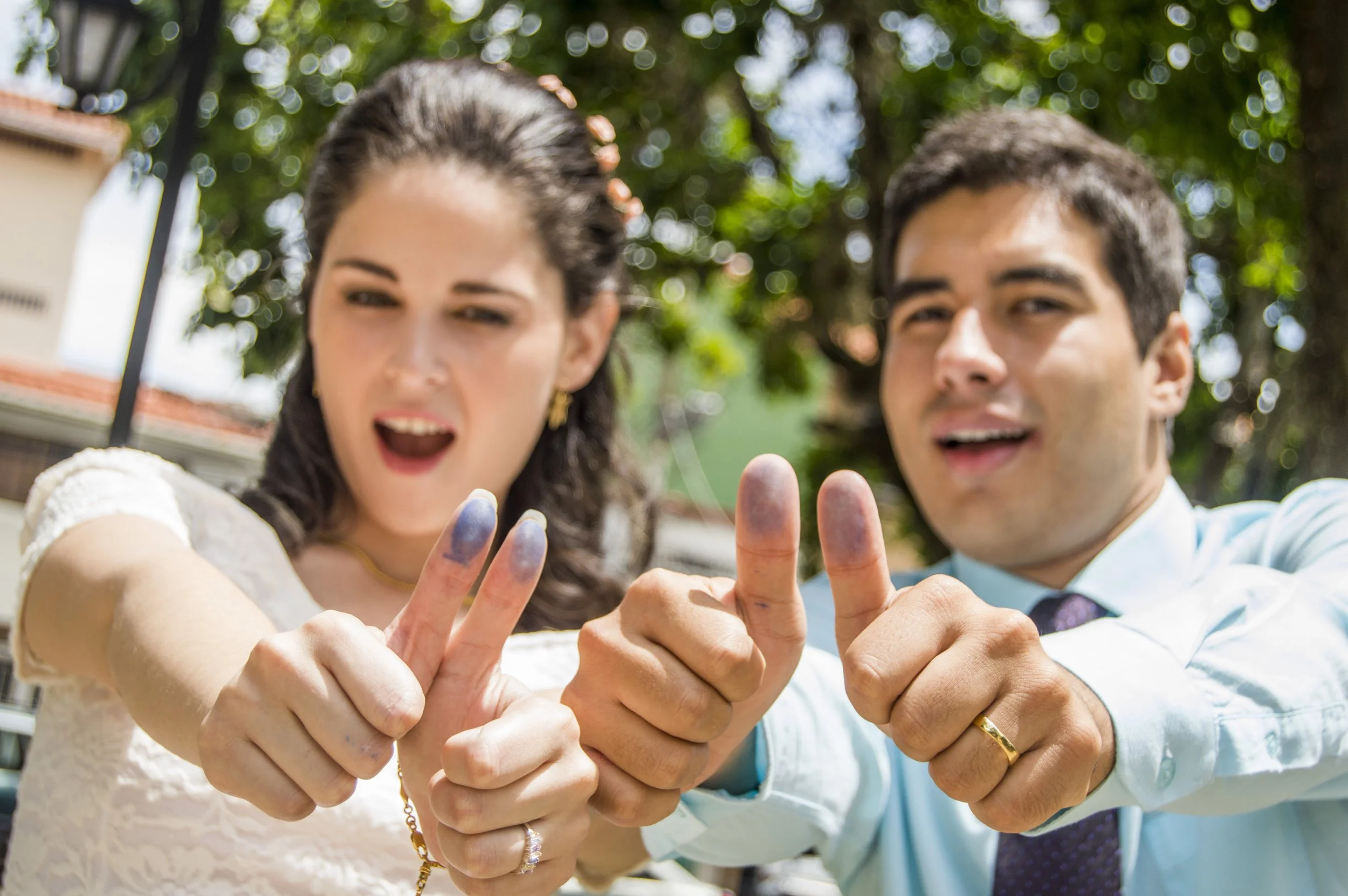 A man and woman are smiling as they pose together, giving thumbs up and making peace signs with their hands; they are outdoors with green trees in the background.