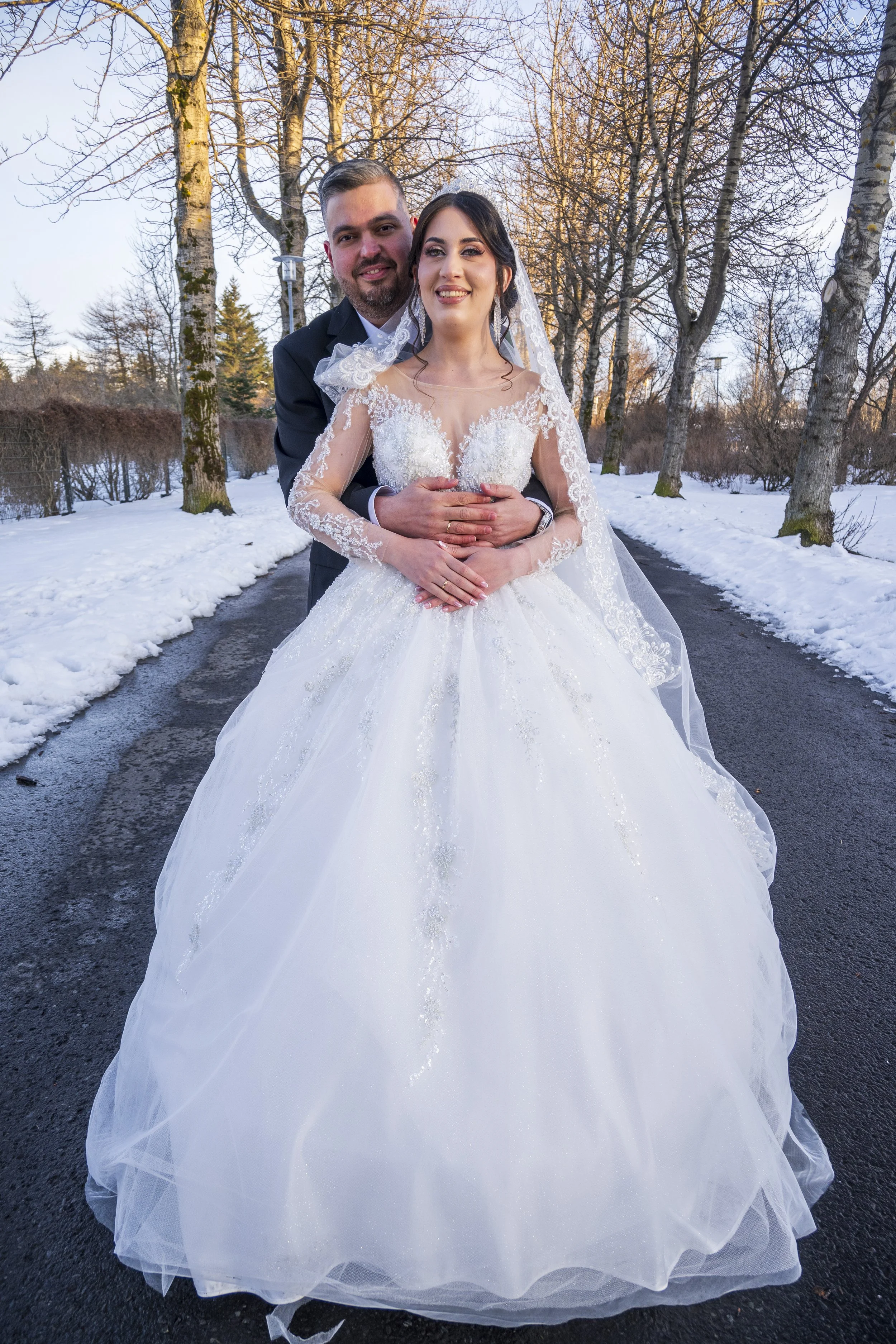A newlywed couple in a winter outdoor setting, the bride in a white wedding gown with lace details and veil, the groom in a dark suit, standing on a snow-covered path with leafless trees in the background.