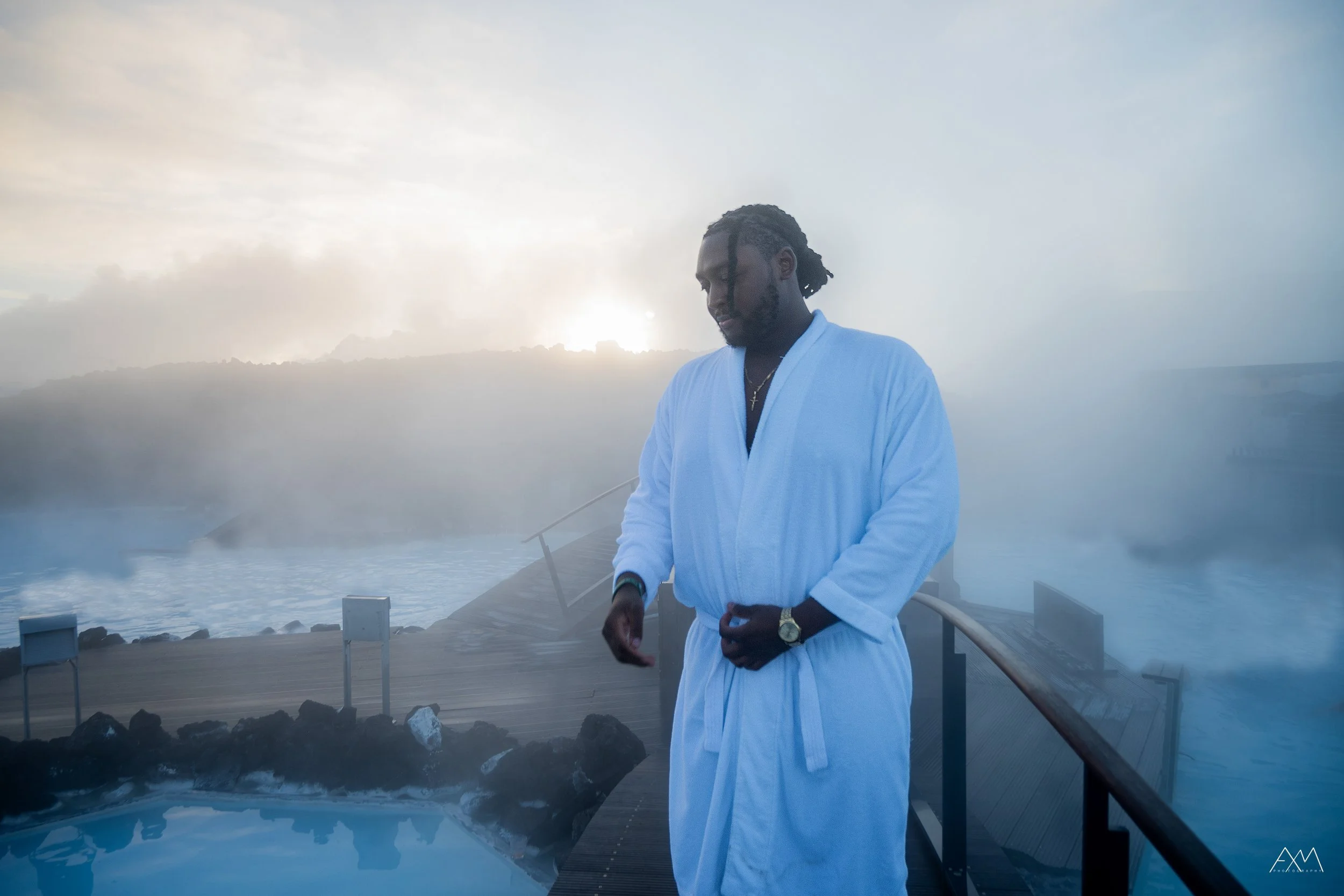 A man in a white bathrobe stands on a wooden deck near a hot spring, with steam rising around him and mountains in the background at sunrise or sunset.