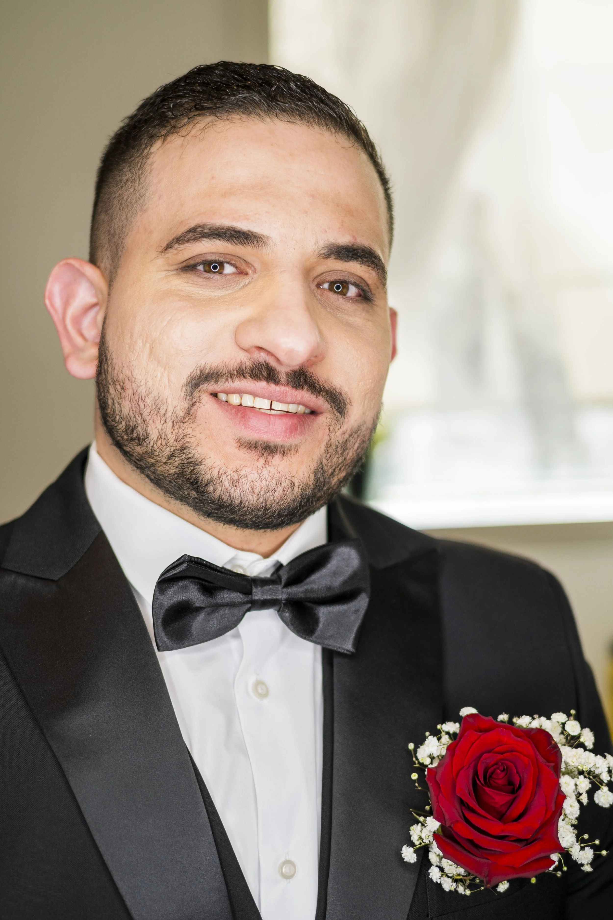A man in a tuxedo with a black bow tie, holding a bouquet of red roses with white baby's breath, smiling.