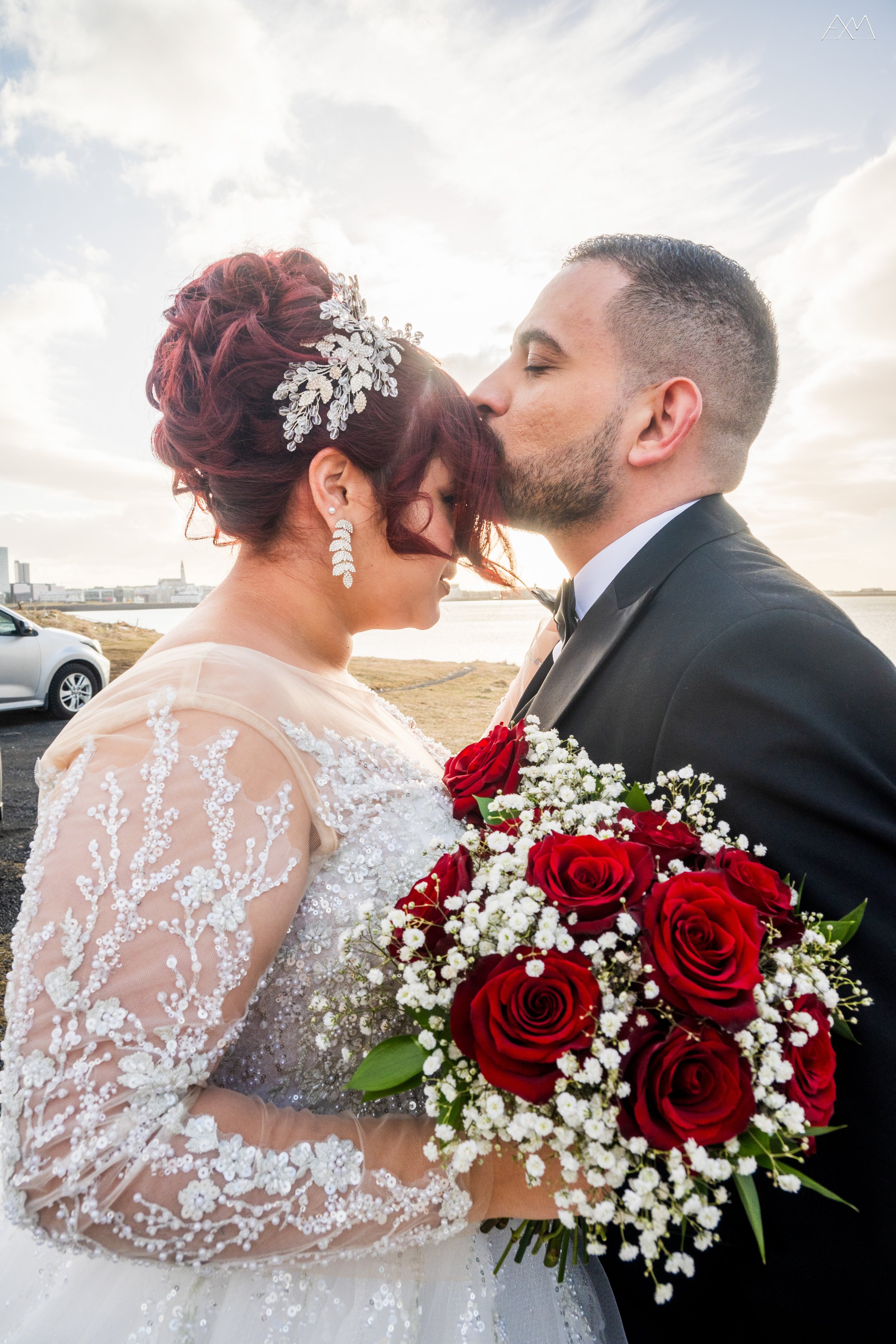 A bride and groom share a kiss outdoors near the water during sunset. The bride wears a lace wedding dress and a decorative hairpiece, holding a bouquet of red roses and white baby's breath. The groom is dressed in a black tuxedo.