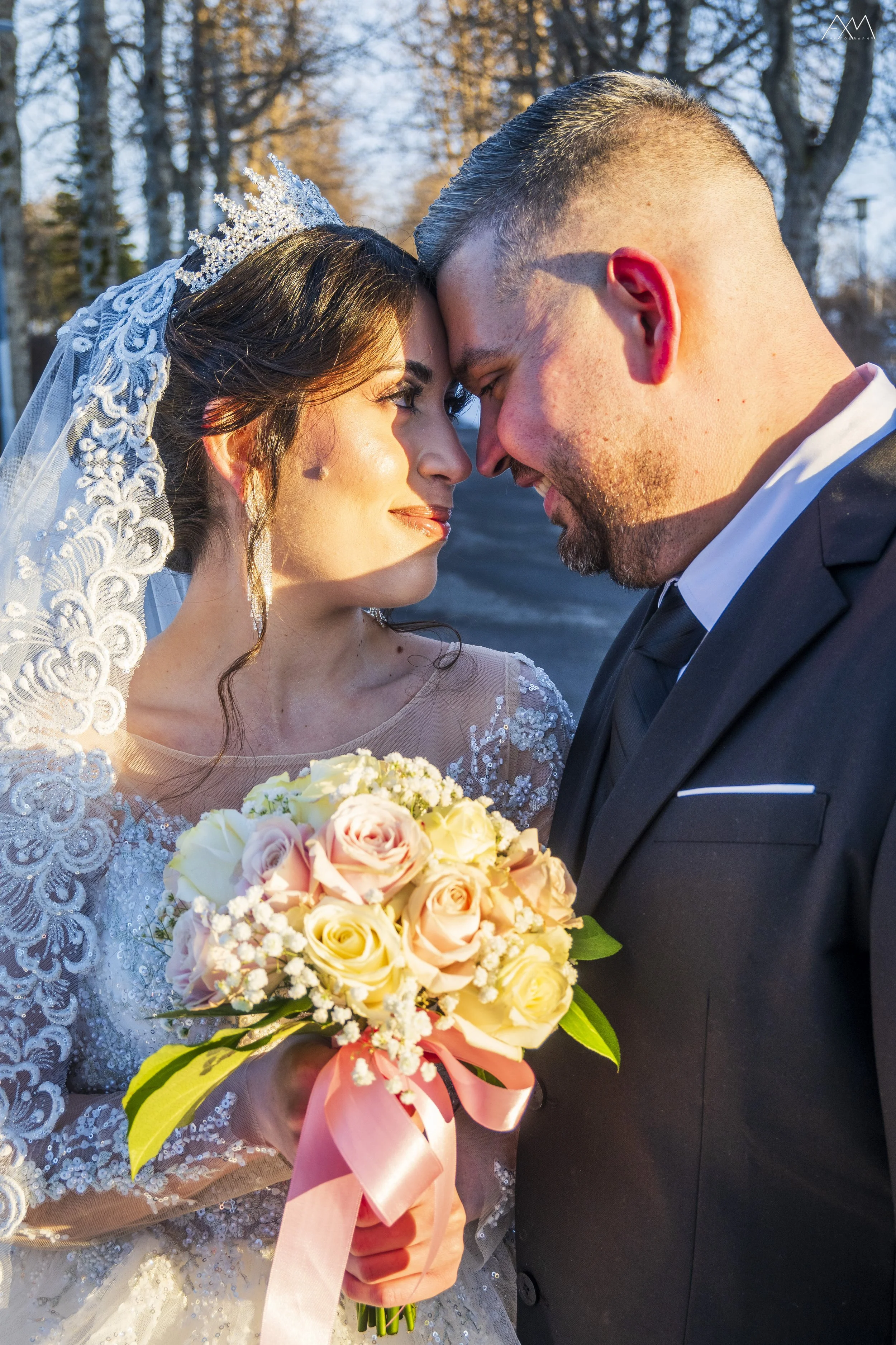 A bride and groom face to face, touching foreheads during their wedding ceremony. The bride is holding a large bouquet of pale yellow, pink, and white roses tied with a pink ribbon. She wears a lace wedding dress with intricate details and a crown wi