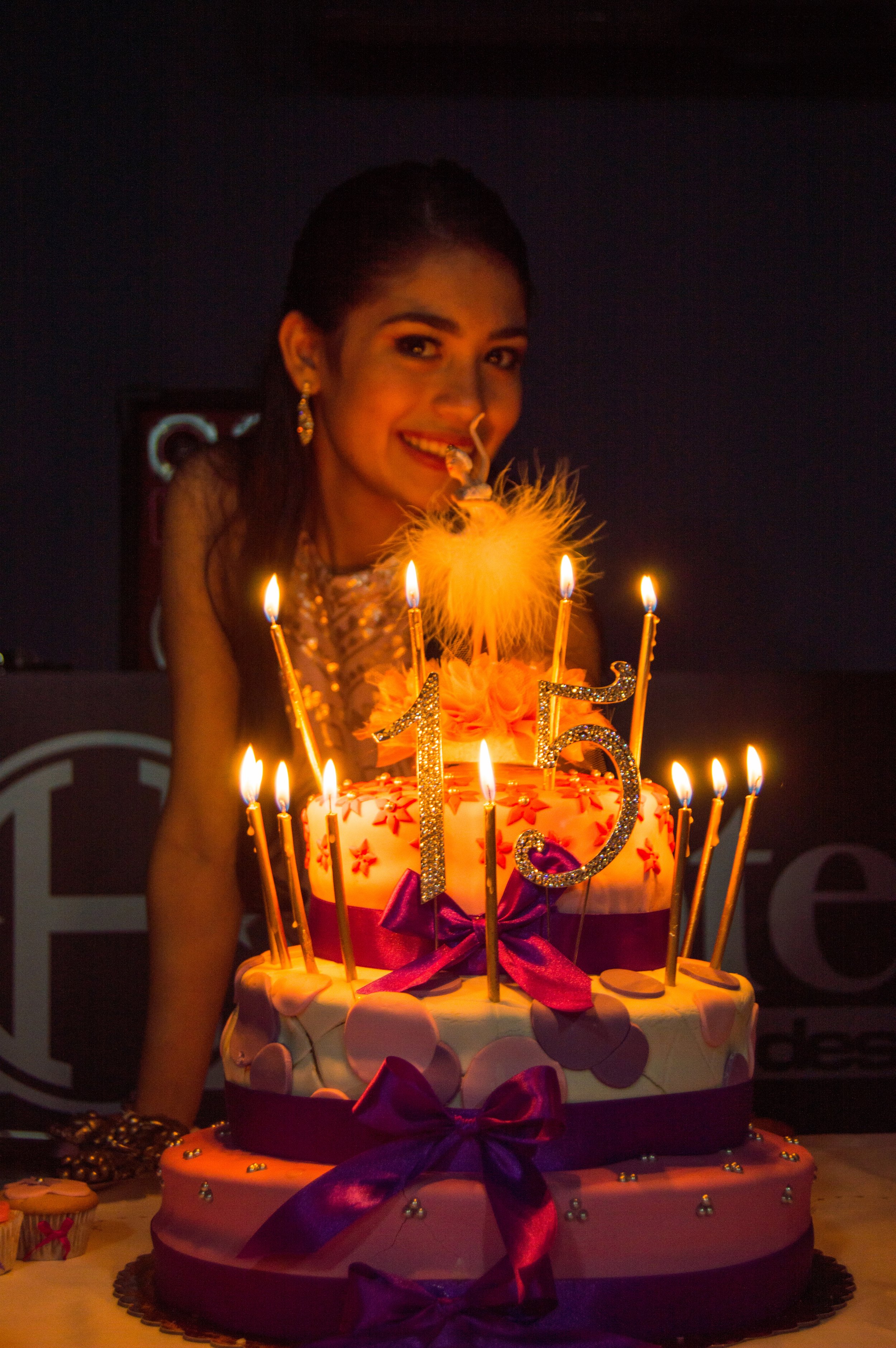 Young woman smiling at birthday celebration with a three-tiered cake decorated with purple ribbons, pink flowers, and lit candles, including a '15' candle, in a dark room with a blue background.