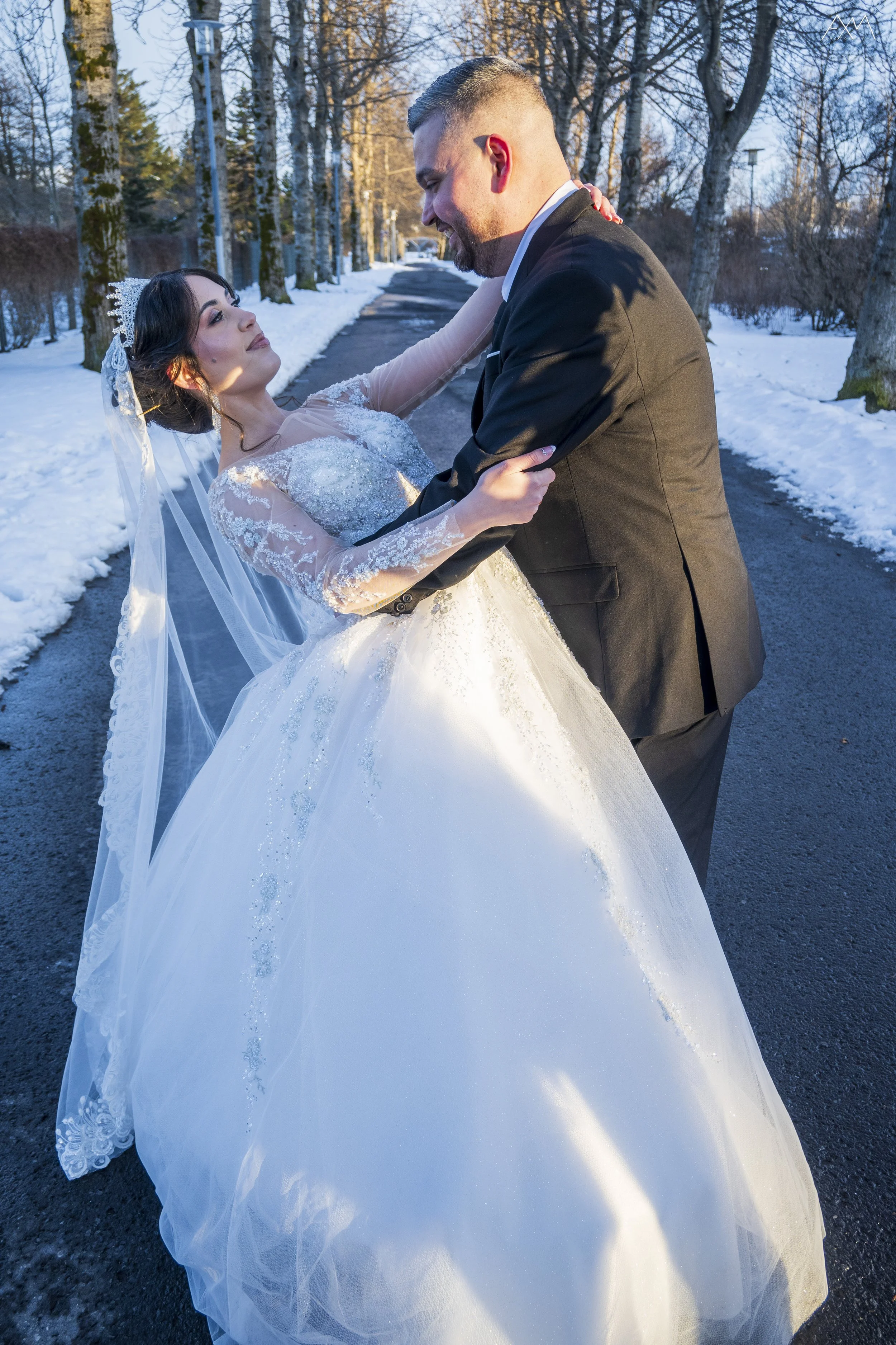 A bride and groom dancing outdoors on a snowy path surrounded by leafless trees during winter.
