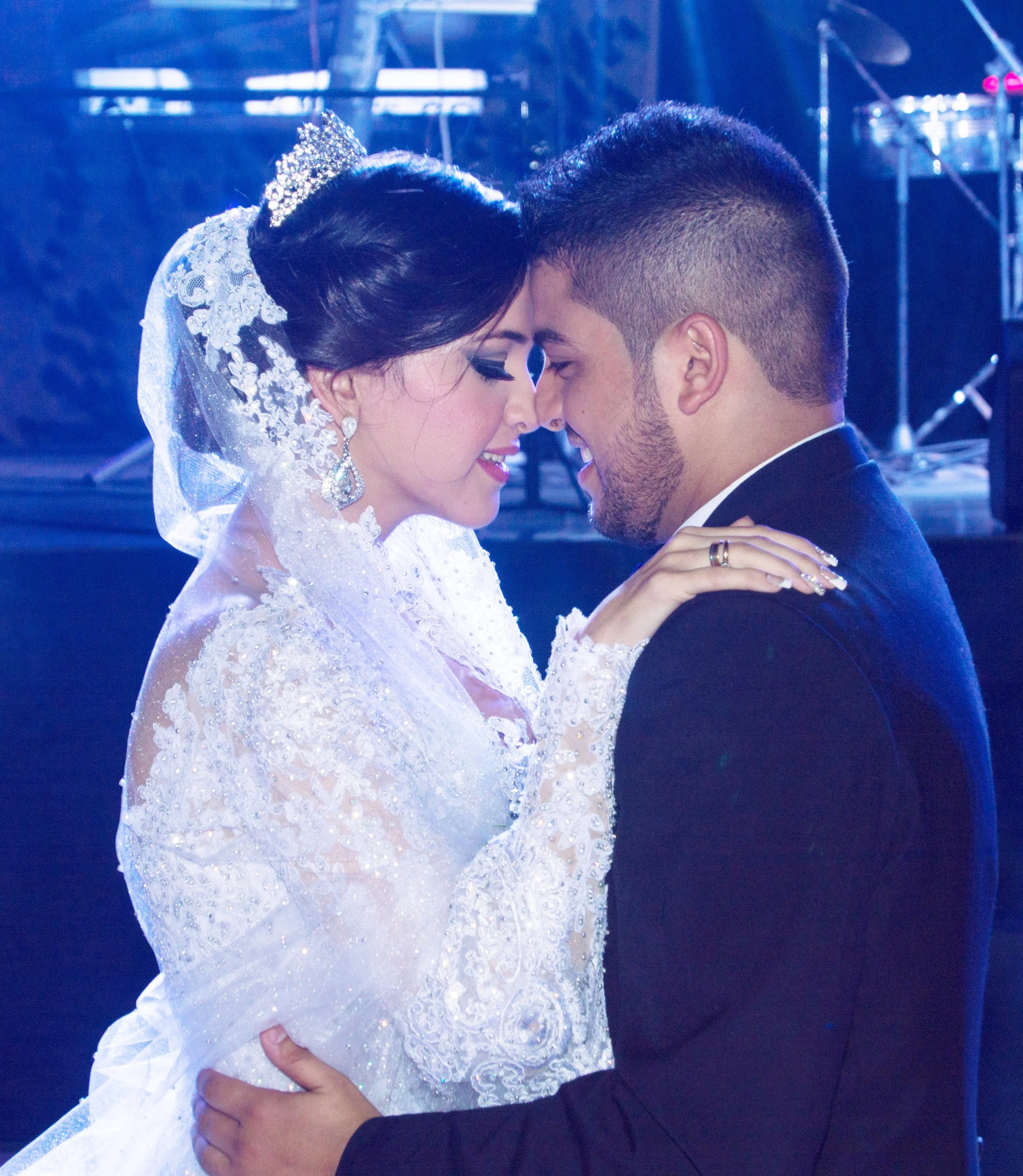 A bride and groom dance closely, touching foreheads and smiling, in a wedding celebration setting with blue lighting.