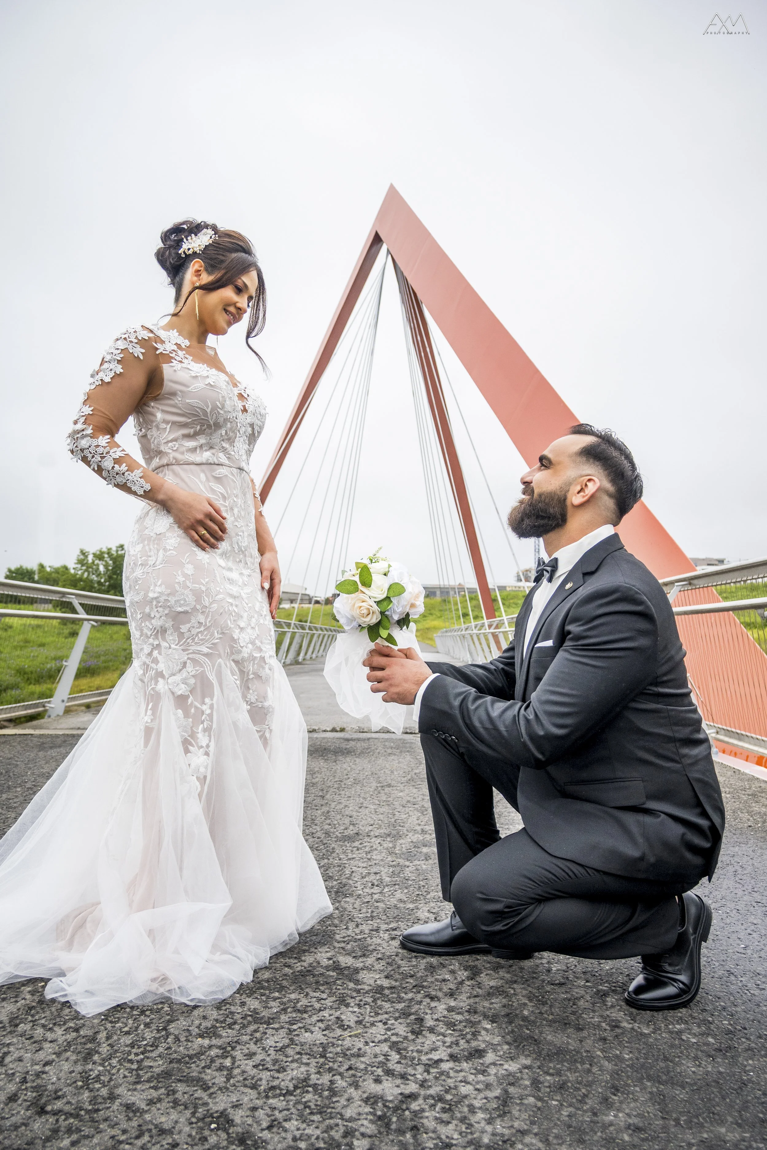 A bride in a white lace wedding dress with long sleeves looking down at her groom, who is kneeling in front of her and proposing with a bouquet of white roses, on a bridge with a red modern arch structure in the background.