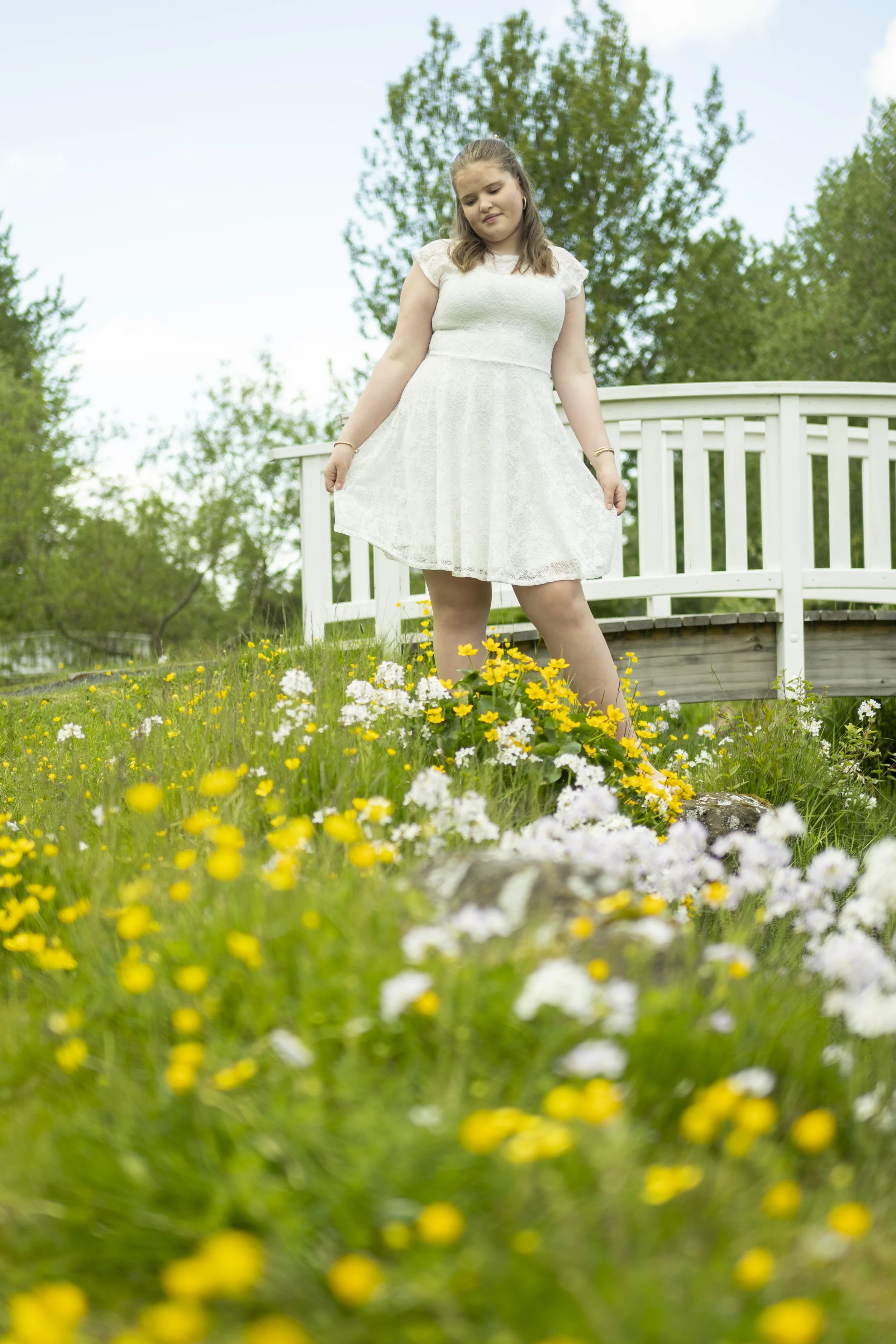 A young woman in a white lace dress standing in a field of yellow and white flowers with a white wooden bridge and green trees in the background.